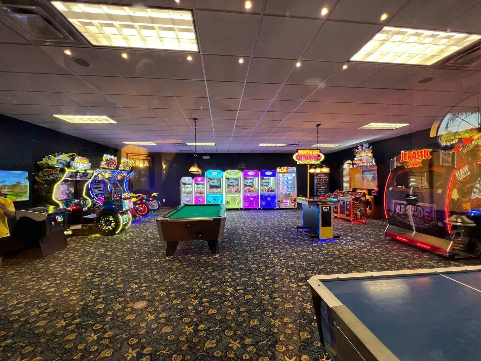 A room with a pool table, arcade games, and a neon sign for Jurassic Park.