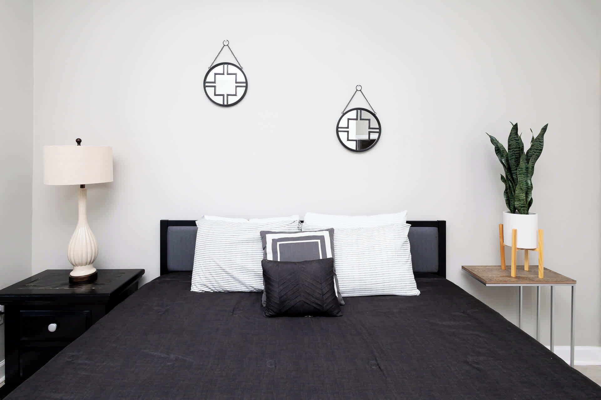 Une chambre à coucher avec un lit noir, des oreillers rayés blancs et gris, un coussin noir, une table de chevet avec une lampe et une plante en pot sur une petite table d’appoint.