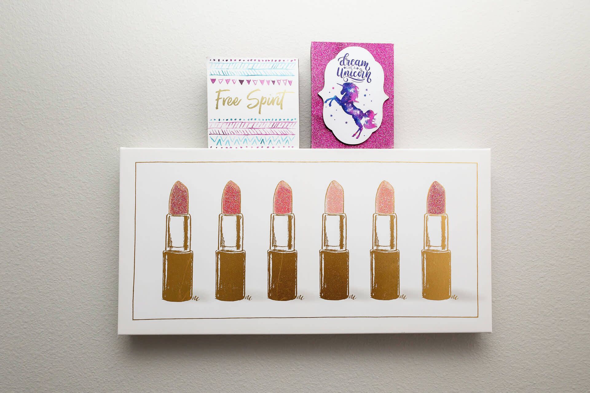 A framed artwork of six lipsticks with glittery tops, a card with 'Free Spirit' text, and a glittery unicorn sticker.