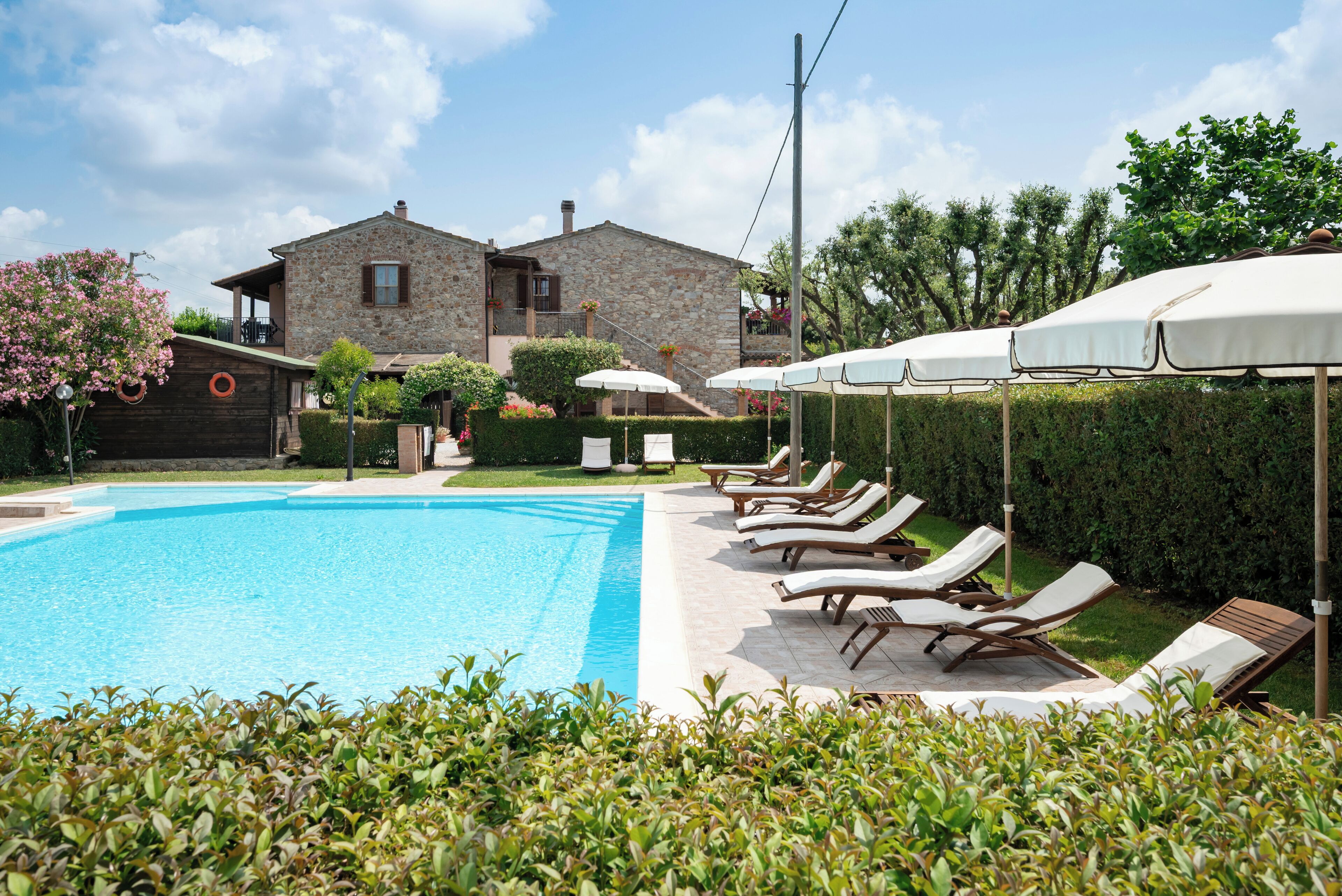 Ein Swimmingpool mit Liegestühlen und Sonnenschirmen, umgeben von Hecken und einem Steinhaus im Hintergrund.