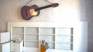 Een kamer met een wit hoofdeinde, een klein nachtkastje met een lamp, een gitaar aan de muur en een raam met jaloezieën.