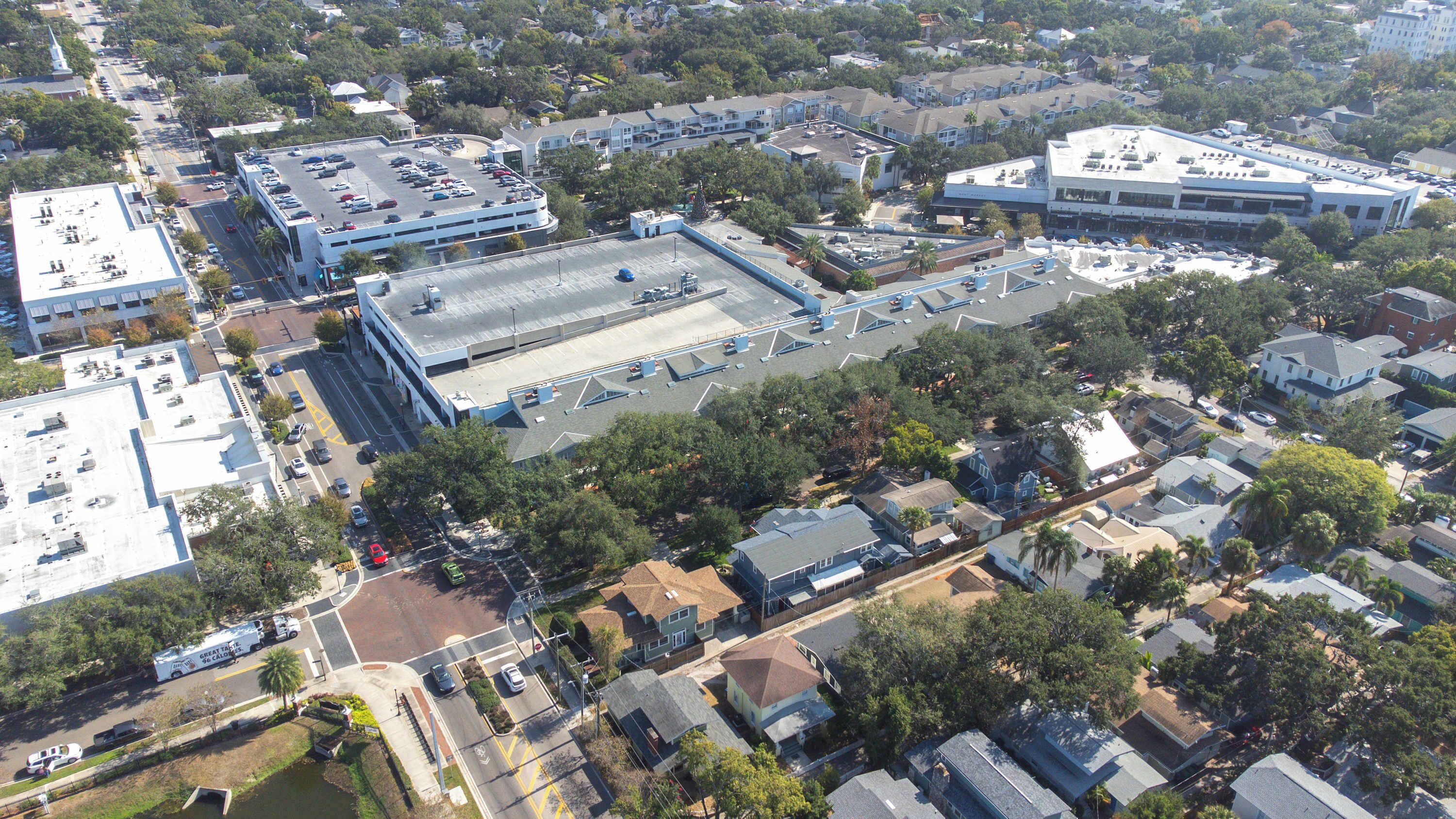 Vue aérienne d’une zone commerciale comprenant un grand parking, un immeuble à plusieurs étages doté d’un toit plat et des maisons résidentielles environnantes.