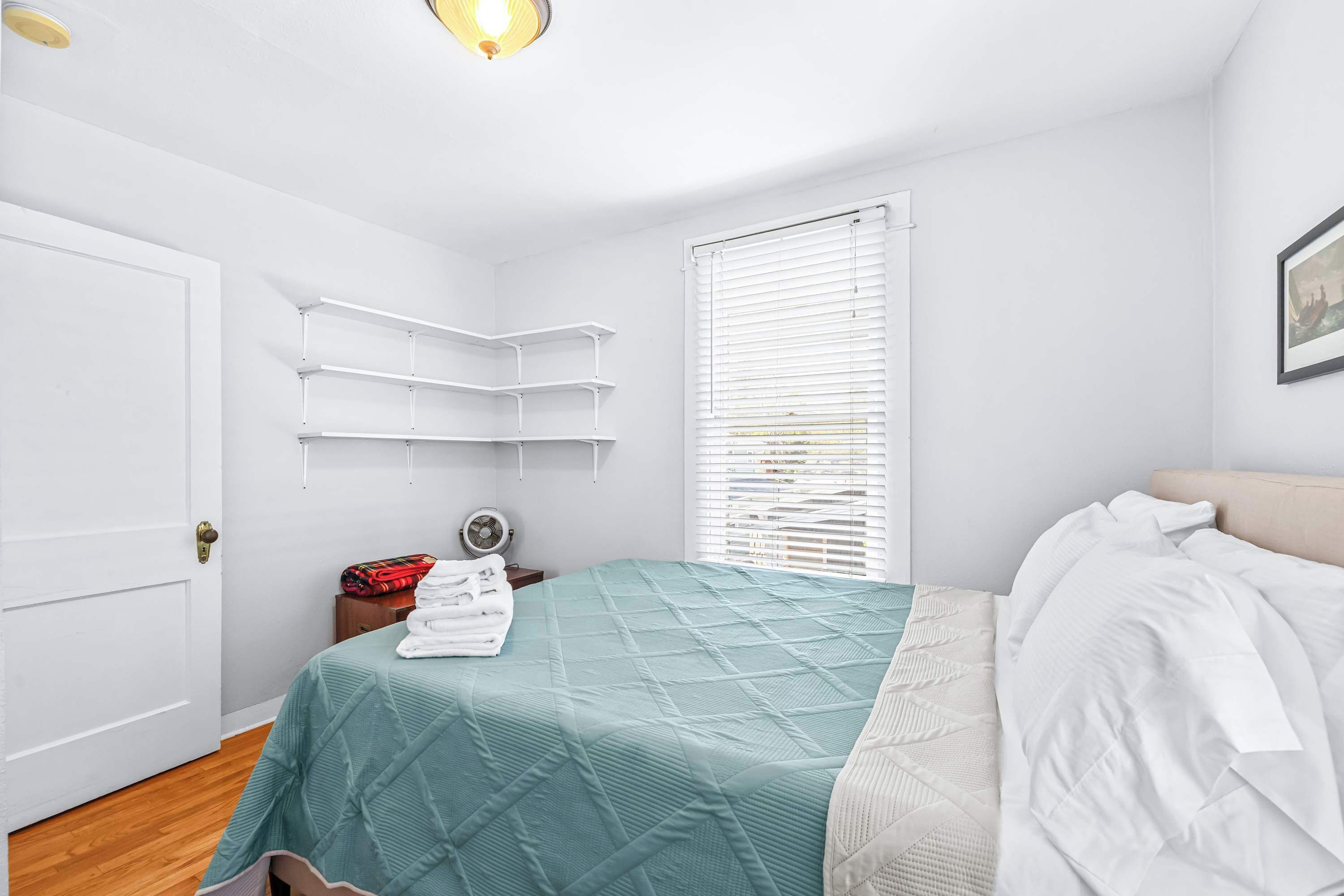 A bedroom with a teal bedspread, white pillows, a white headboard, and a window with blinds.