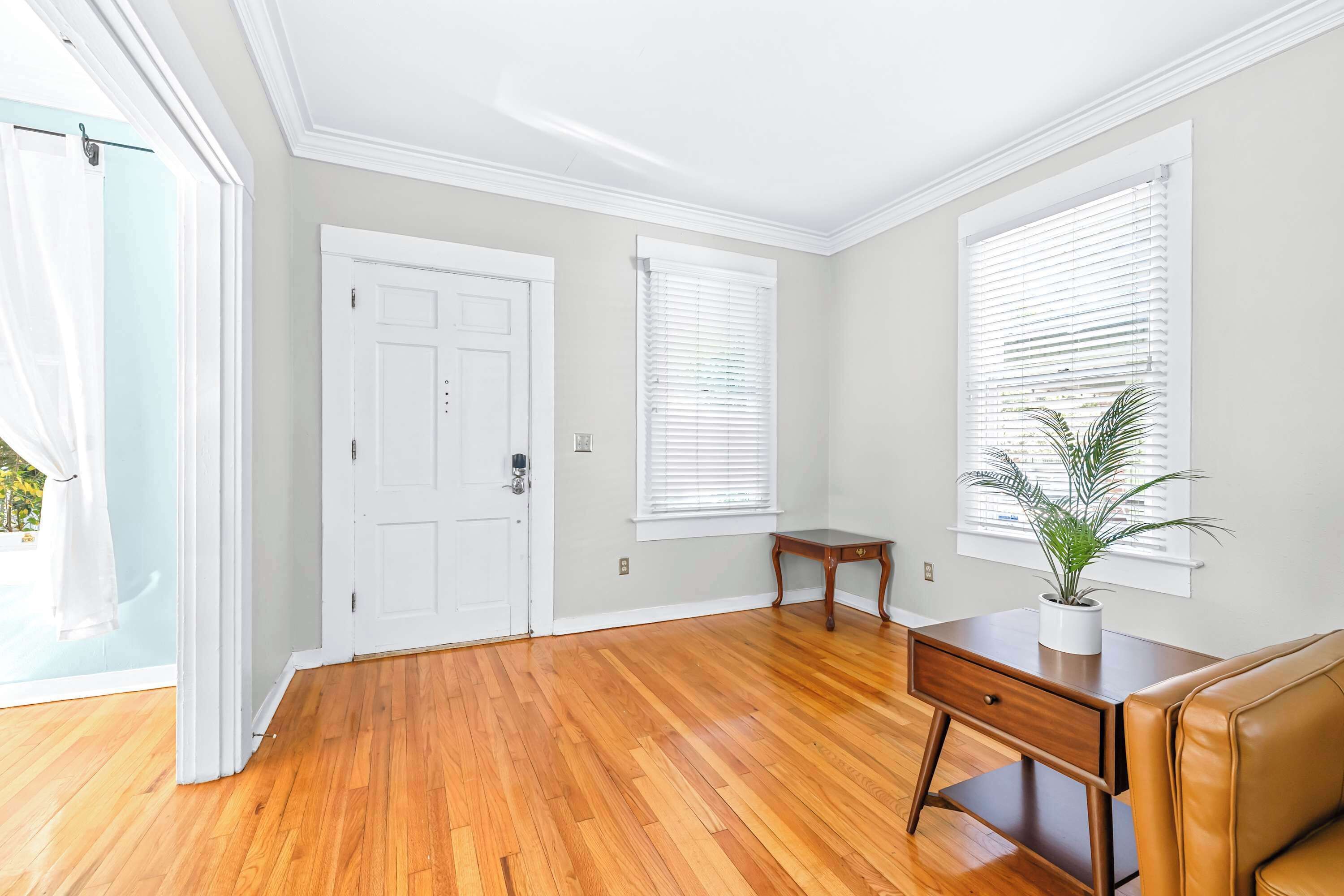 A spacious room with hardwood flooring, a white door, two windows with blinds, a small wooden table, and a brown leather couch.