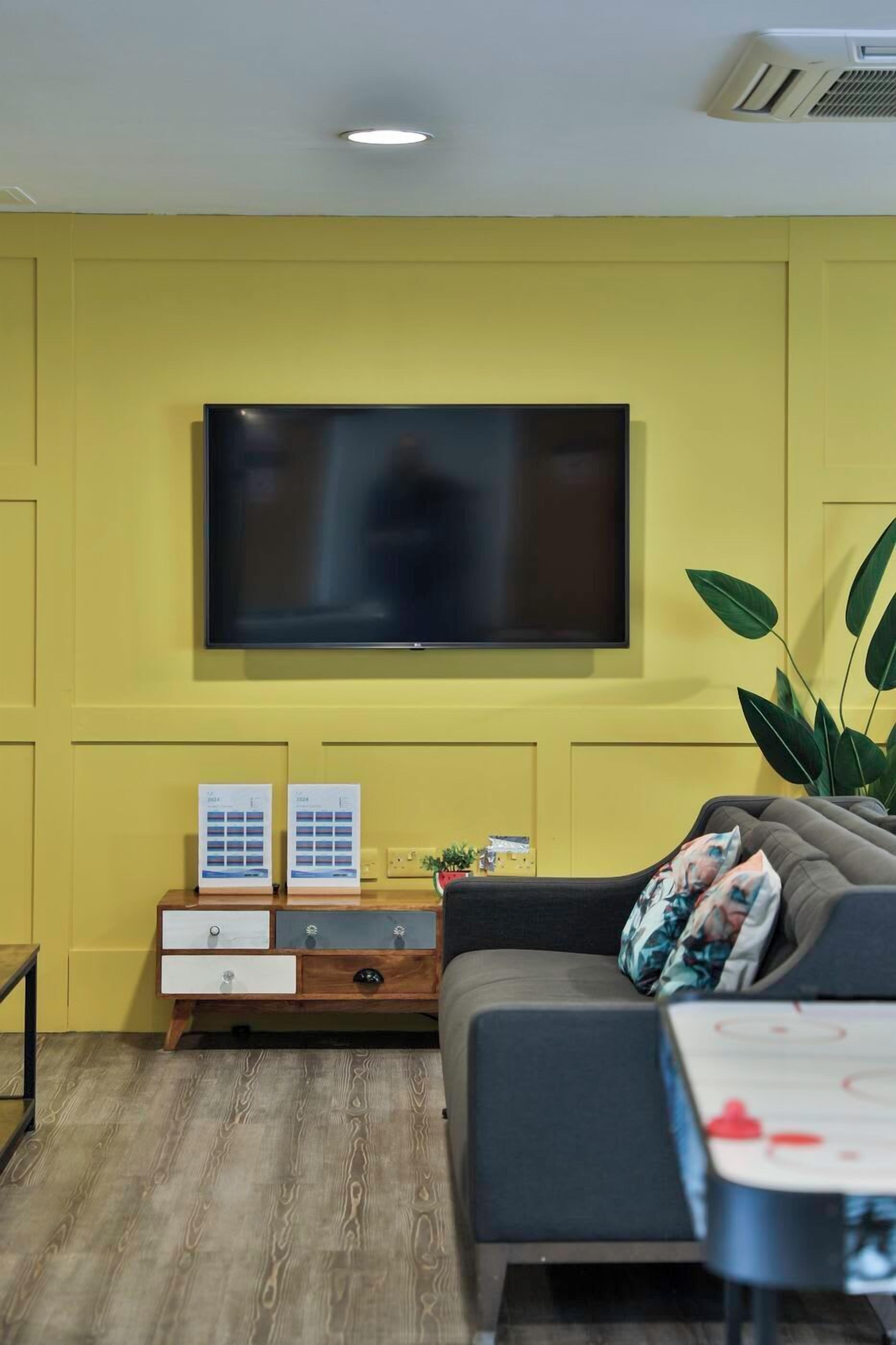 A living room with a flat-screen TV mounted on a yellow wall, a grey sofa, a wooden side table with a plant, and a small table with a hockey puck.