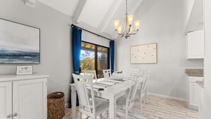 Un comedor con una mesa y sillas blancas, una lámpara de araña, un ventanal con cortinas azules y vistas al exterior.