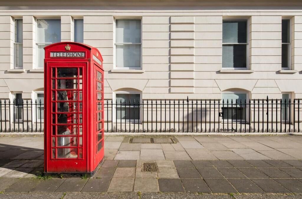 A red British telephone booth in front of a building with windows and a black metal fence.
