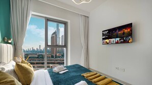 Une chambre d’hôtel avec une grande fenêtre, un lit recouvert de draps blancs, un plaid turquoise et un téléviseur fixé au mur qui diffuse des affiches de films.