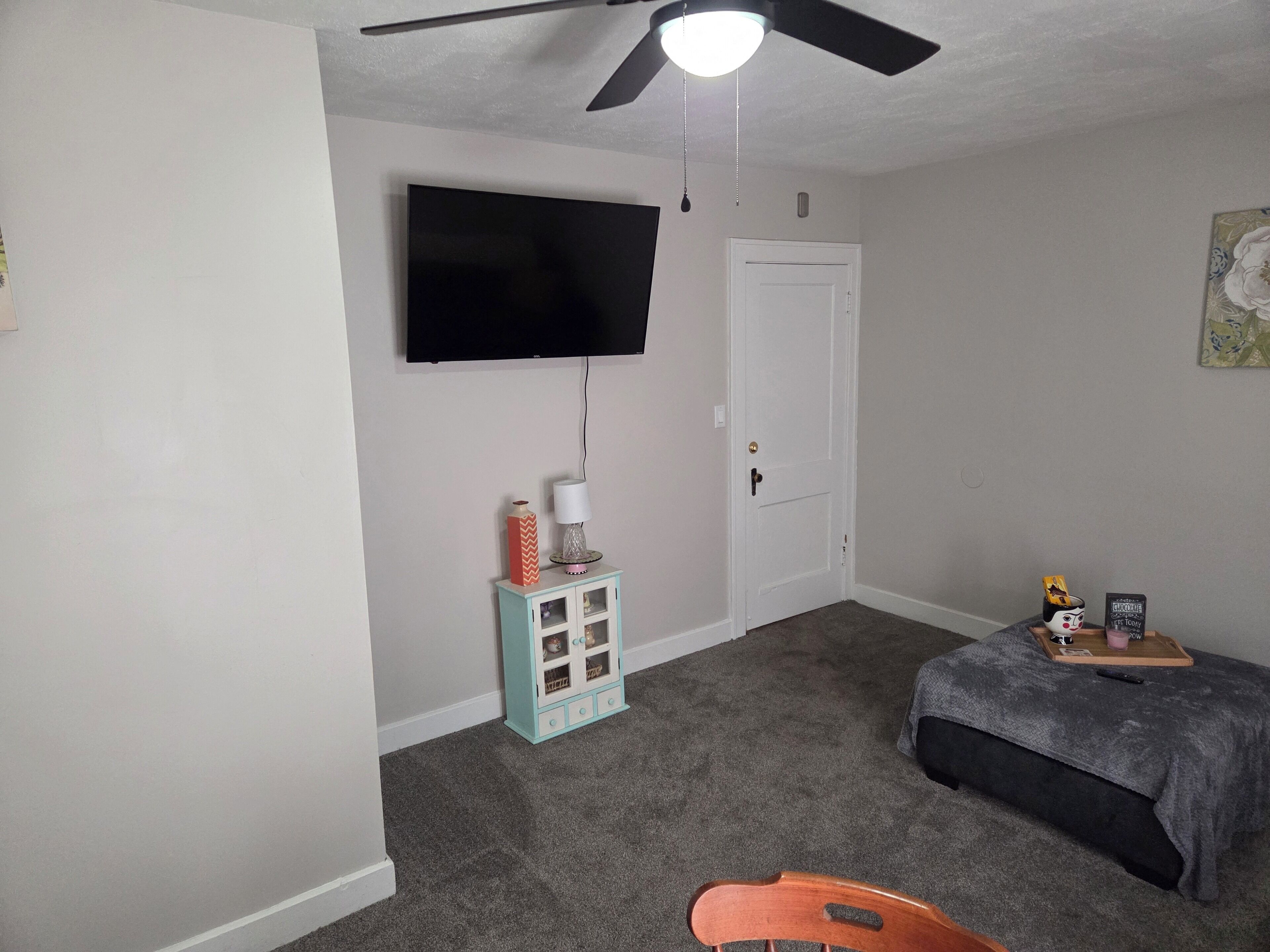 A bedroom with a ceiling fan, a TV mounted on the wall, a door, a small cabinet with decorative items, a bed with a gray blanket, and a chair.