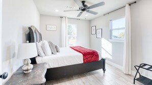 A modern bedroom with a bed, a ceiling fan, a bedside table, and a window. - Modern 4BR Retreat Near Downtown & Trails (Bentonville)