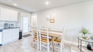 A modern kitchen with white cabinets, a dining table with wooden chairs, and a small side table with a vase. - Modern 4BR Retreat Near Downtown & Trails (Bentonville)