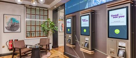 A hotel lobby with check-in kiosks, a seating area with a table and chairs, and a large window with a city view.