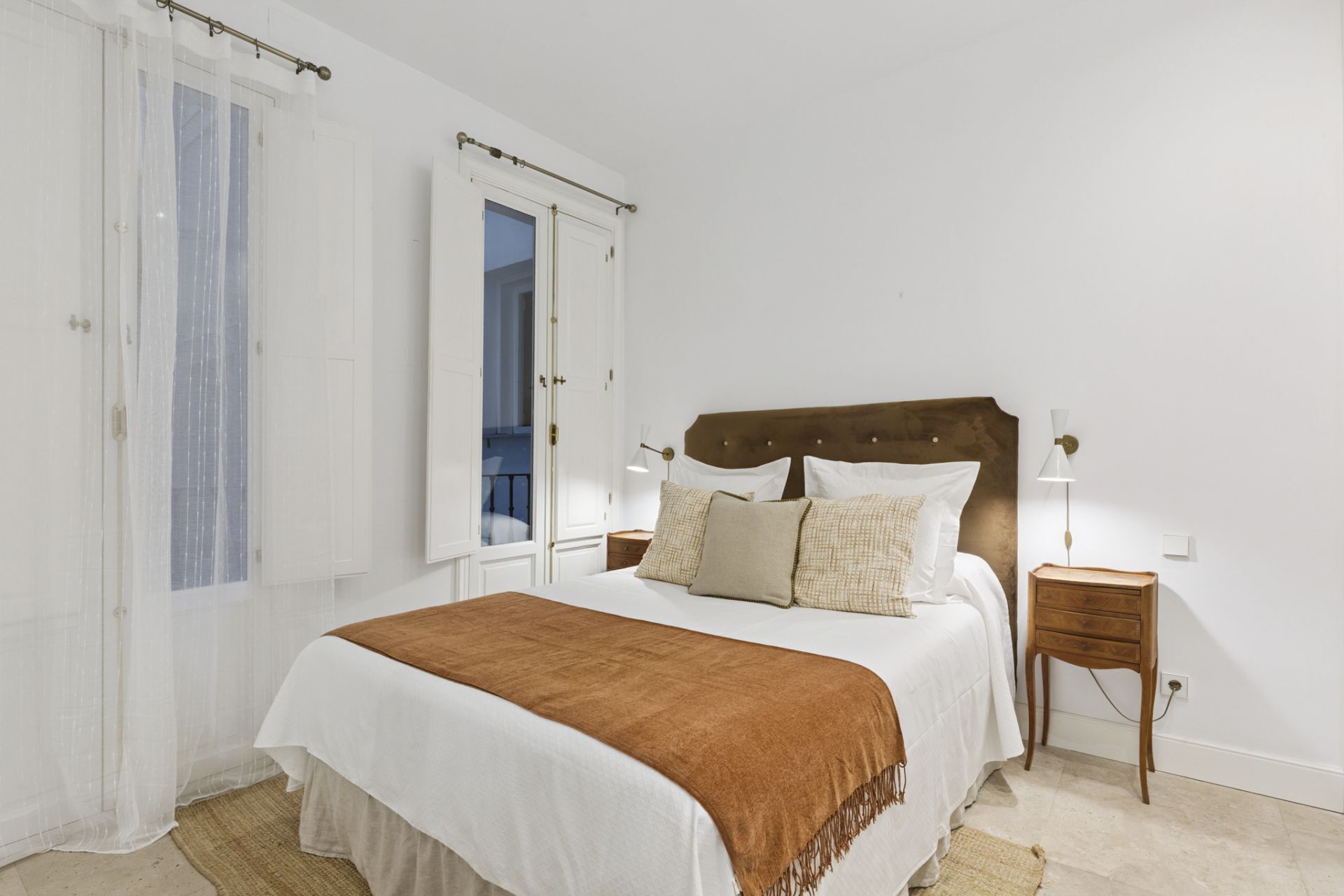 Un dormitorio con cama, dos mesitas de noche, una mesita auxiliar, una ventana con cortinas y una puerta.