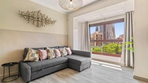 Ein modernes Wohnzimmer mit einer grauen Eckcouch, einem Wandbild und einem großen Fenster mit Blick auf die Stadt.