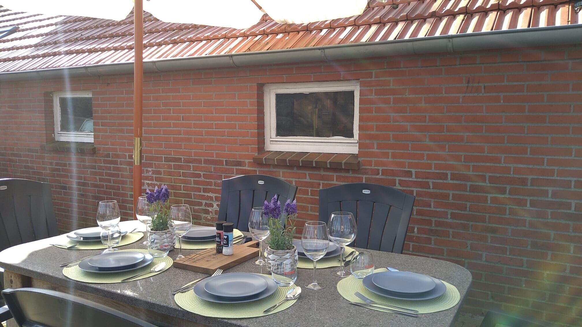 Un espace de restauration en plein air avec un mur en briques, une fenêtre et une table dressée pour quatre personnes, avec des assiettes, des verres et des couverts.