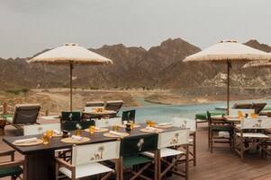 Un área de comedor al aire libre con mesas y sillas, sombrillas y vistas a montañas y un cuerpo de agua.