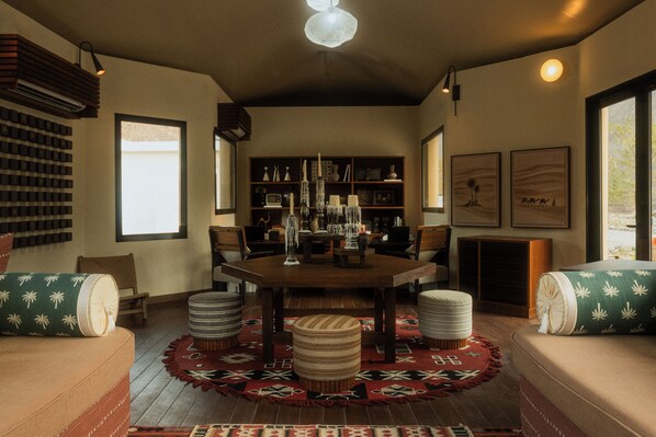 A cozy living room with a wooden dining table, cushioned seating, a patterned rug, and a bookshelf filled with various items. - Caravan Hatta By Our Habitas (Hatta)