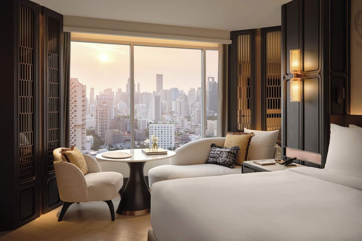 Ein modernes Hotelzimmer mit einem großen Bett, einem runden Tisch, zwei Sesseln und Blick auf eine Stadtkulisse durch raumhohe Fenster.