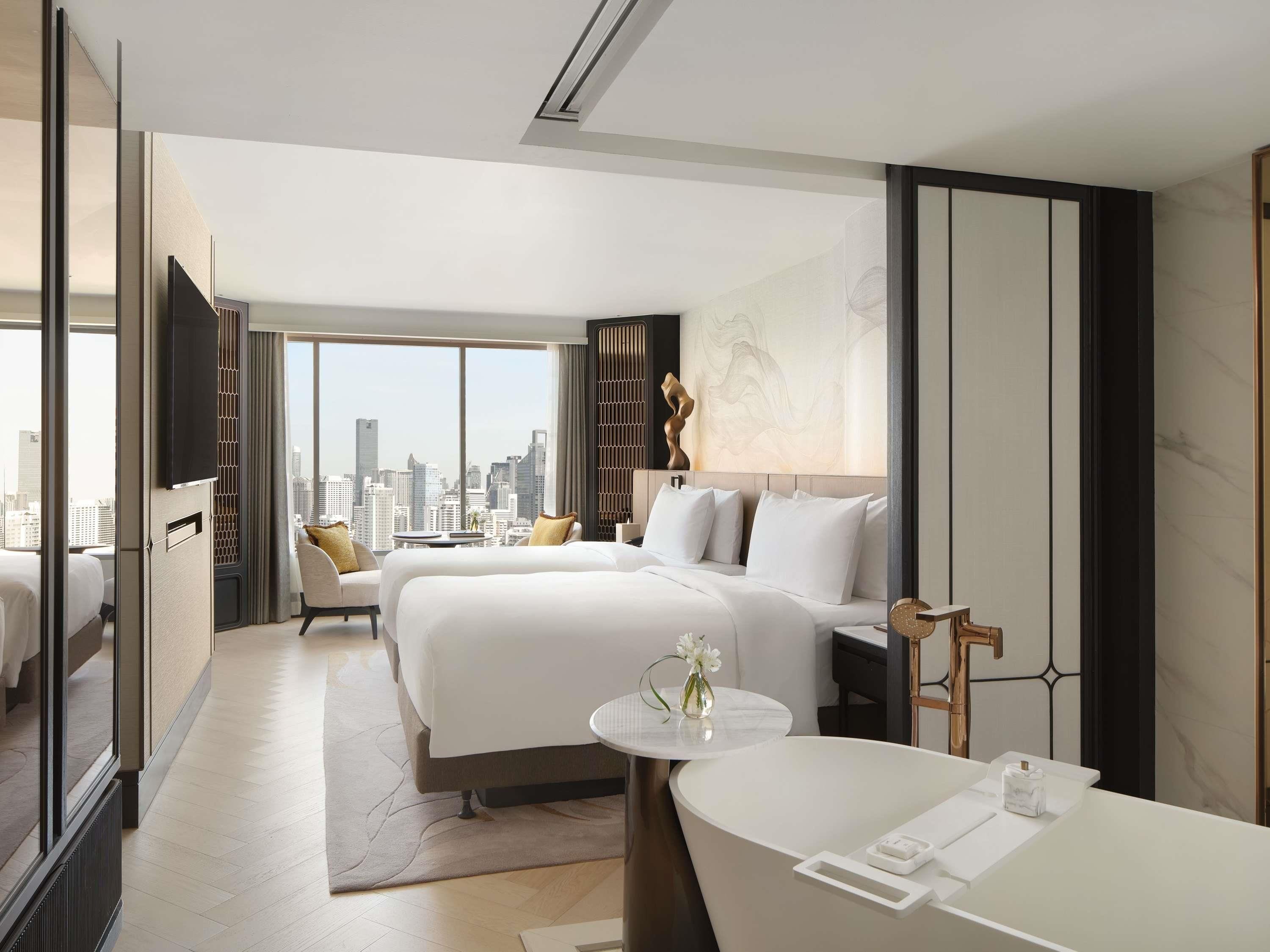 A modern hotel room with a city view, a large bed, a separate seating area, and a bathroom with a bathtub.