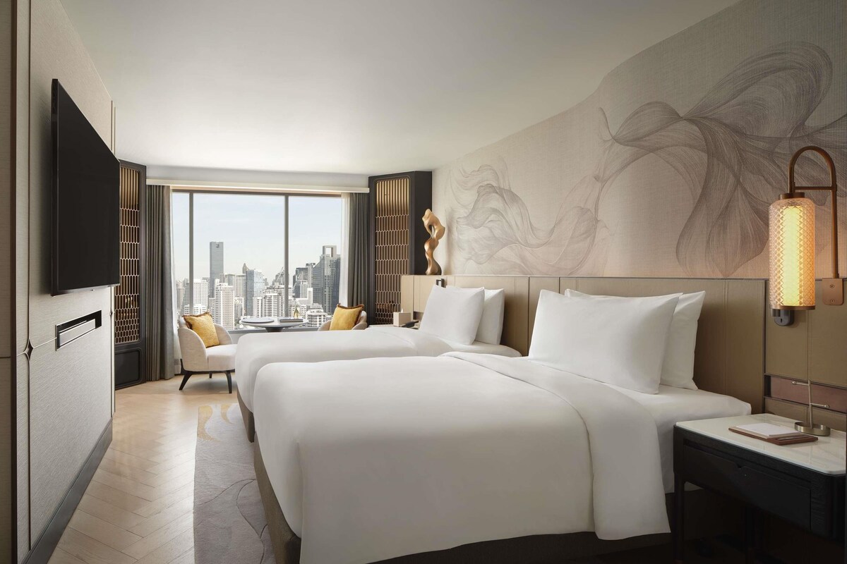 Ein Hotelzimmer mit zwei Betten, einem Schreibtisch, einem Stuhl, einem Fernseher und einem großen Fenster mit Blick auf die Stadt.
