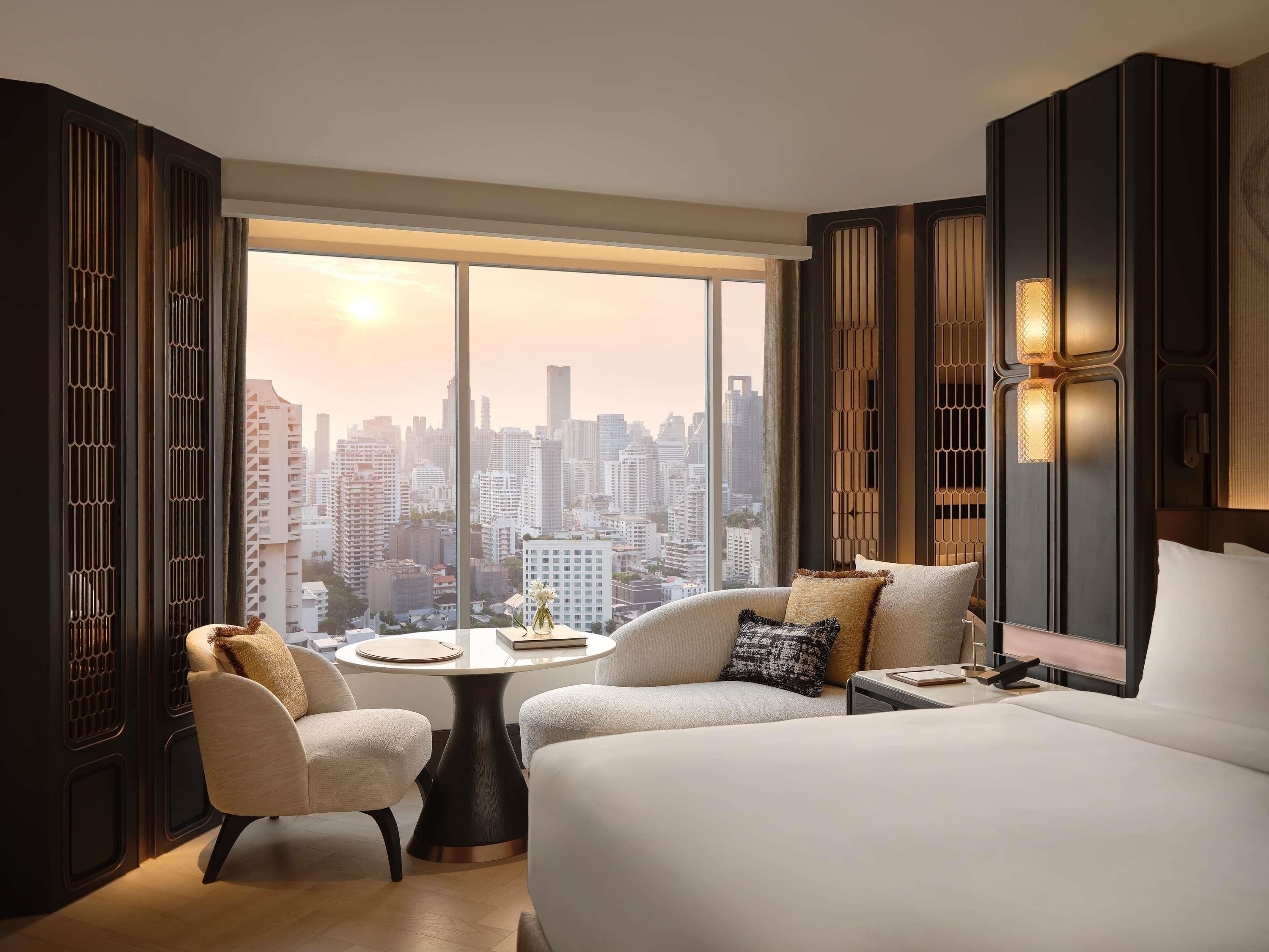 A modern hotel room with a large bed, a round table, two armchairs, and a view of a cityscape.