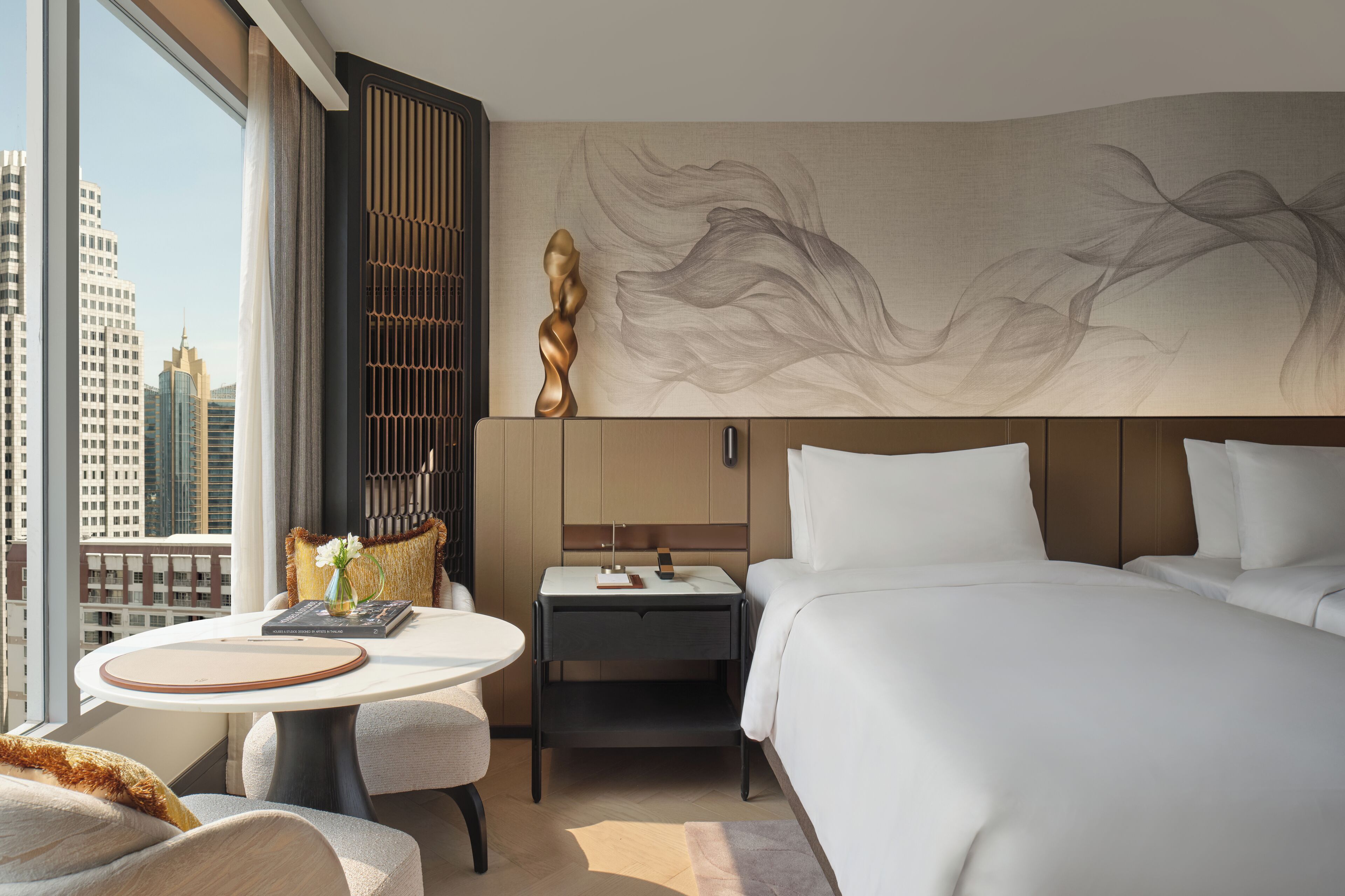 A hotel room with a large bed, a bedside table, a round table, a view of the cityscape, and a decorative wall piece.