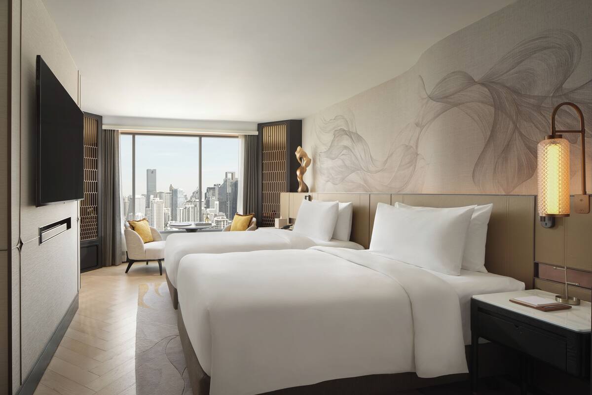Ein Hotelzimmer mit zwei Betten, einem Schreibtisch, einem Sessel und einem Fernseher, mit Blick auf die Stadt.
