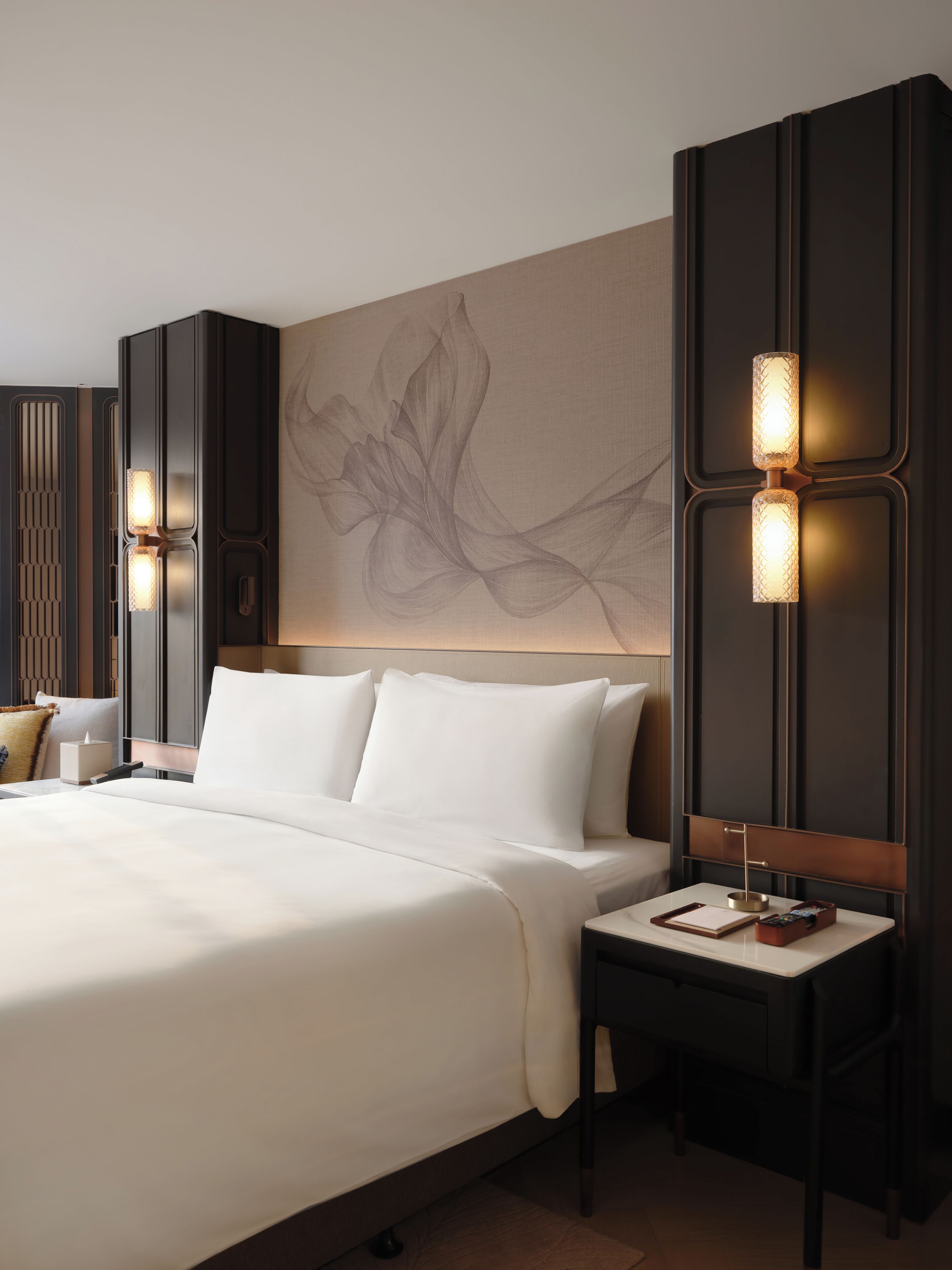 A modern hotel room with a large bed, a nightstand with a lamp, and a decorative wall piece.