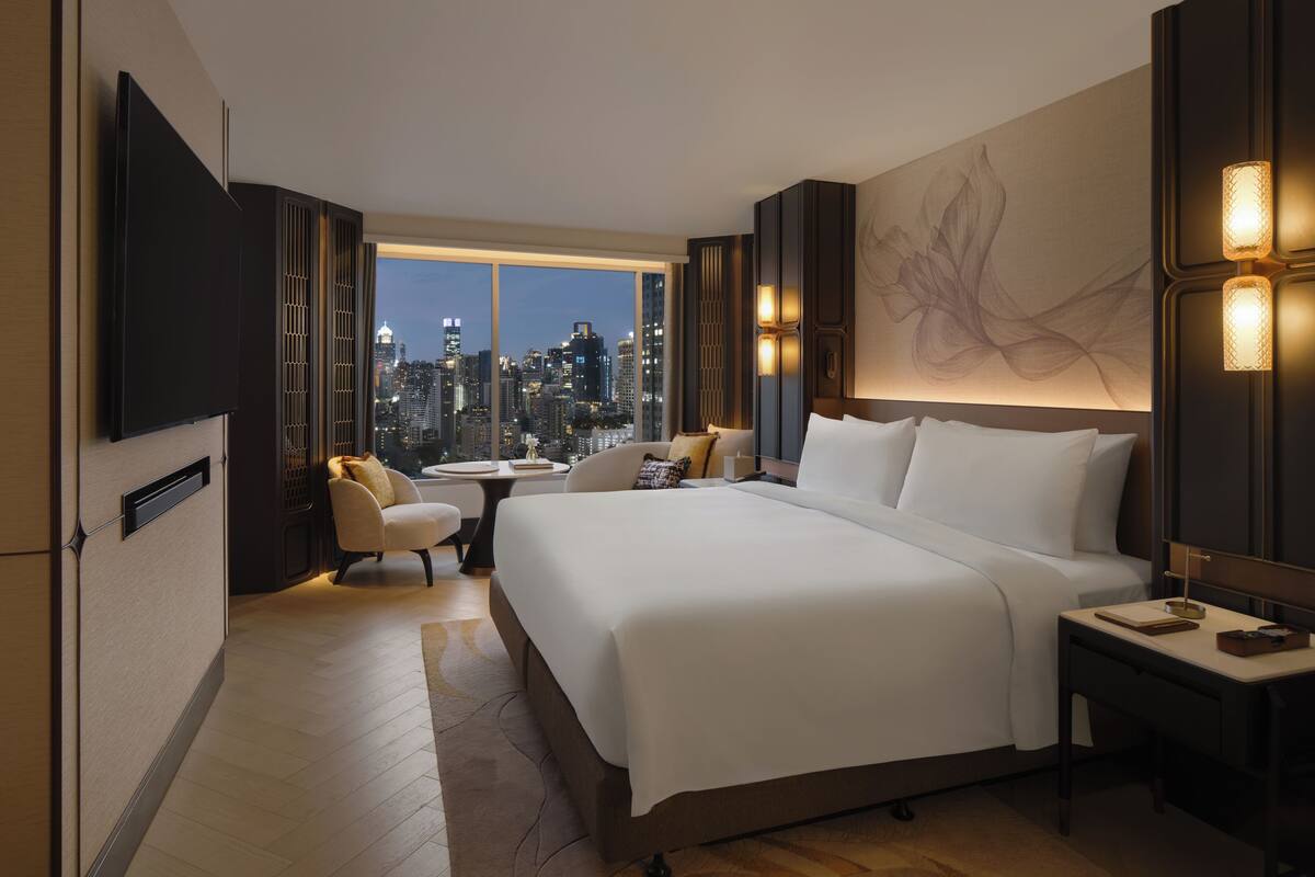 Ein Hotelzimmer mit einem großen Bett, einem Fernseher, Blick auf die Stadt und einem Schreibtisch mit Lampe.