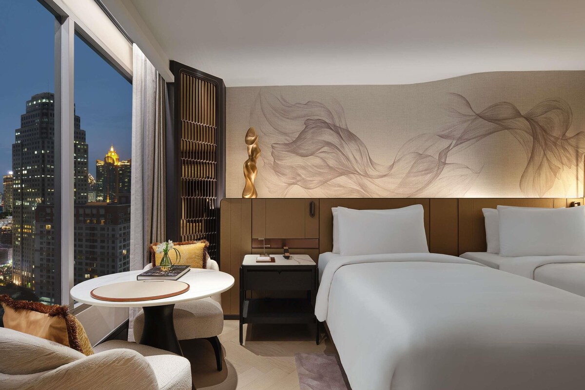 A hotel room with a large bed, a bedside table, a chair, a desk, and a view of the cityscape.