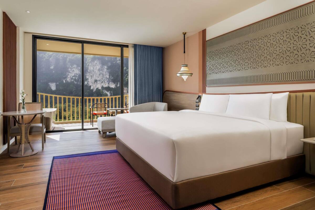 A hotel room with a large bed, a balcony with a view, a dining area, and a decorative wall.