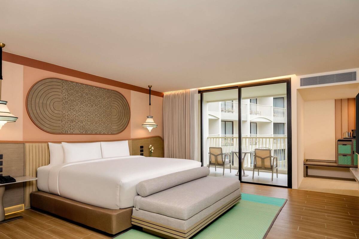 A hotel room with a large bed, a small sofa, a desk, and a balcony with chairs.