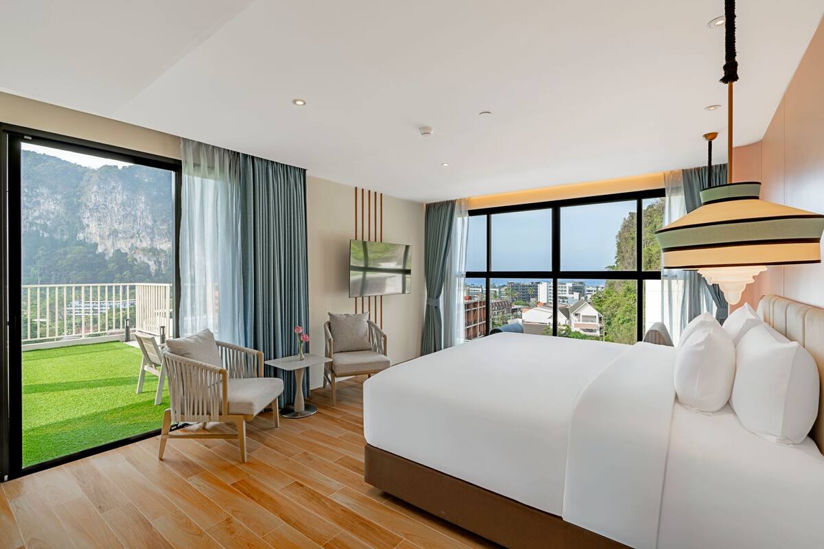 A modern hotel room with a large bed, a balcony with a view of the mountains, a seating area with chairs, and a TV.