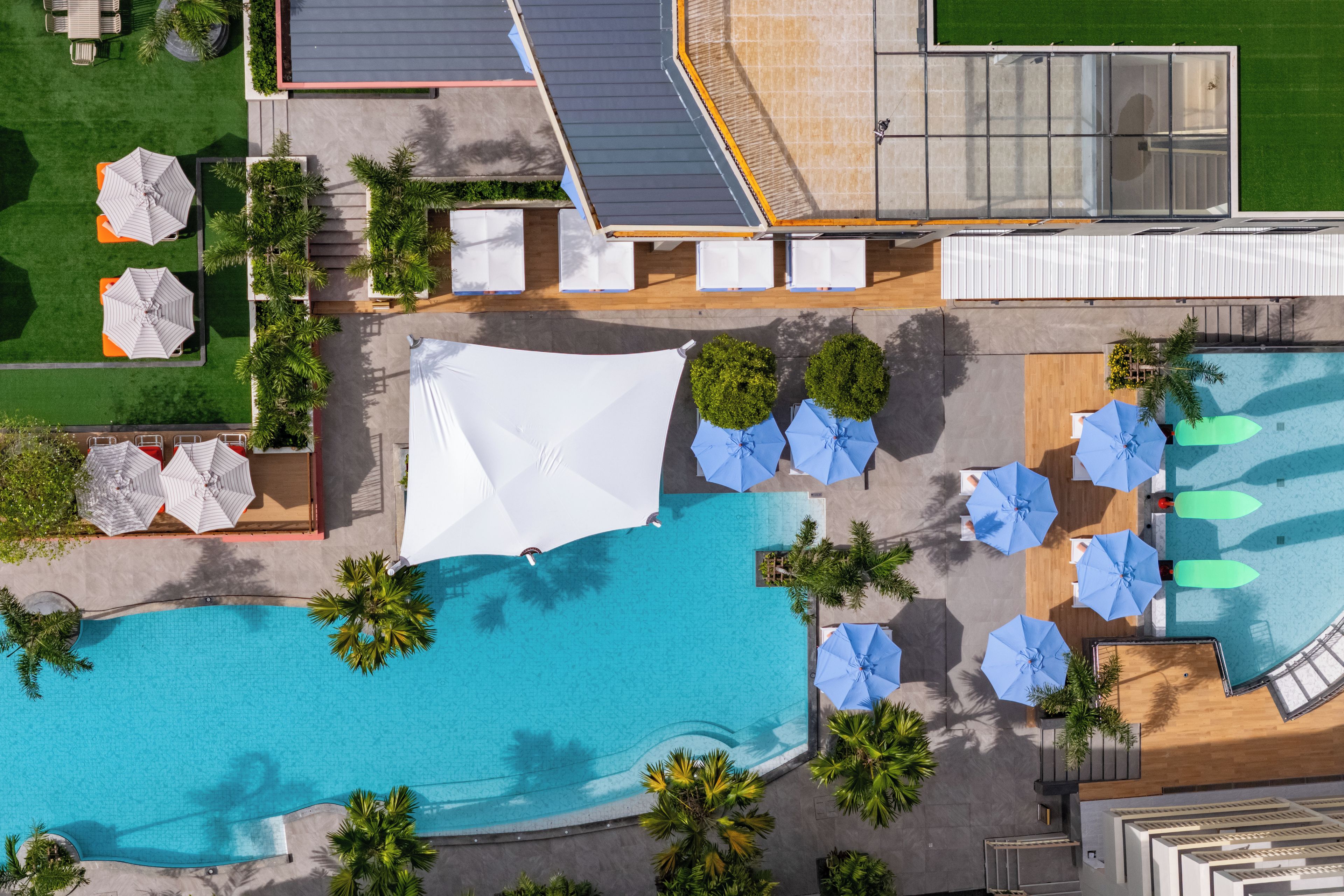 An aerial view of a resort with a swimming pool, sun umbrellas, lounge chairs, and a covered seating area.