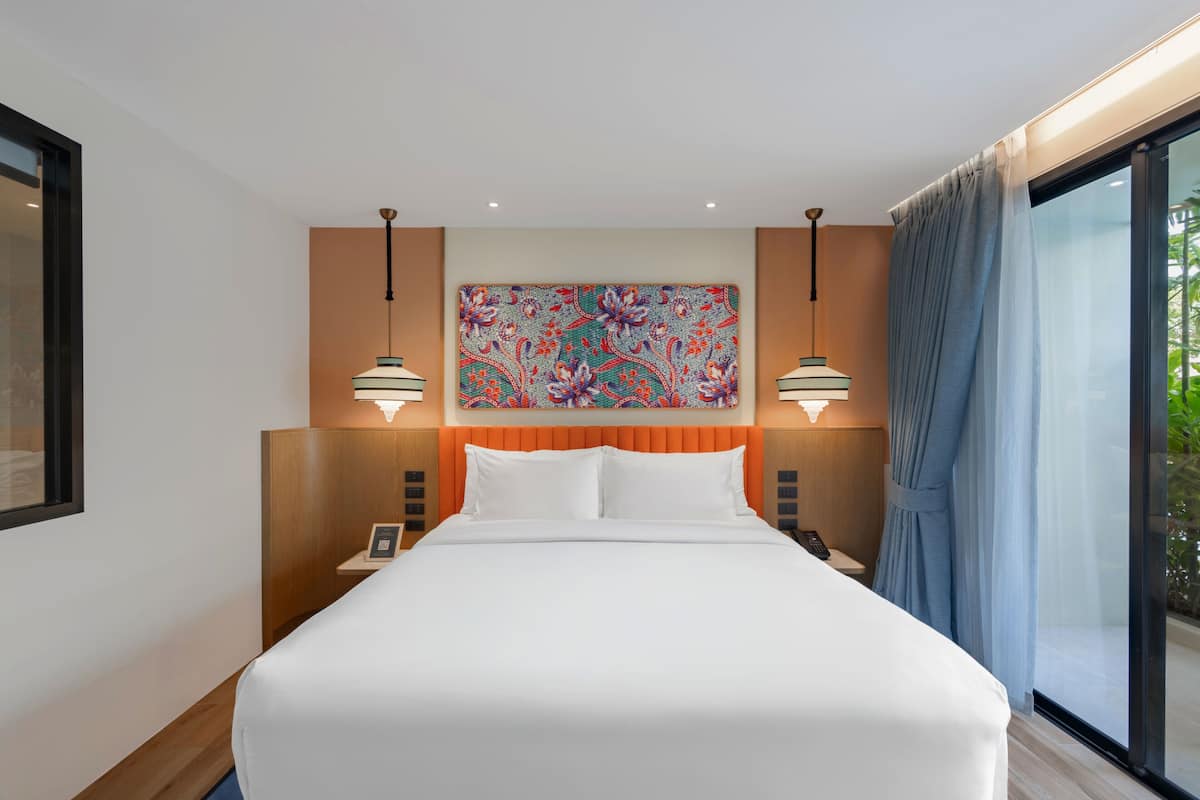 A hotel room with a large bed, two bedside tables with lamps, a colorful abstract painting on the wall, and a large window with curtains.