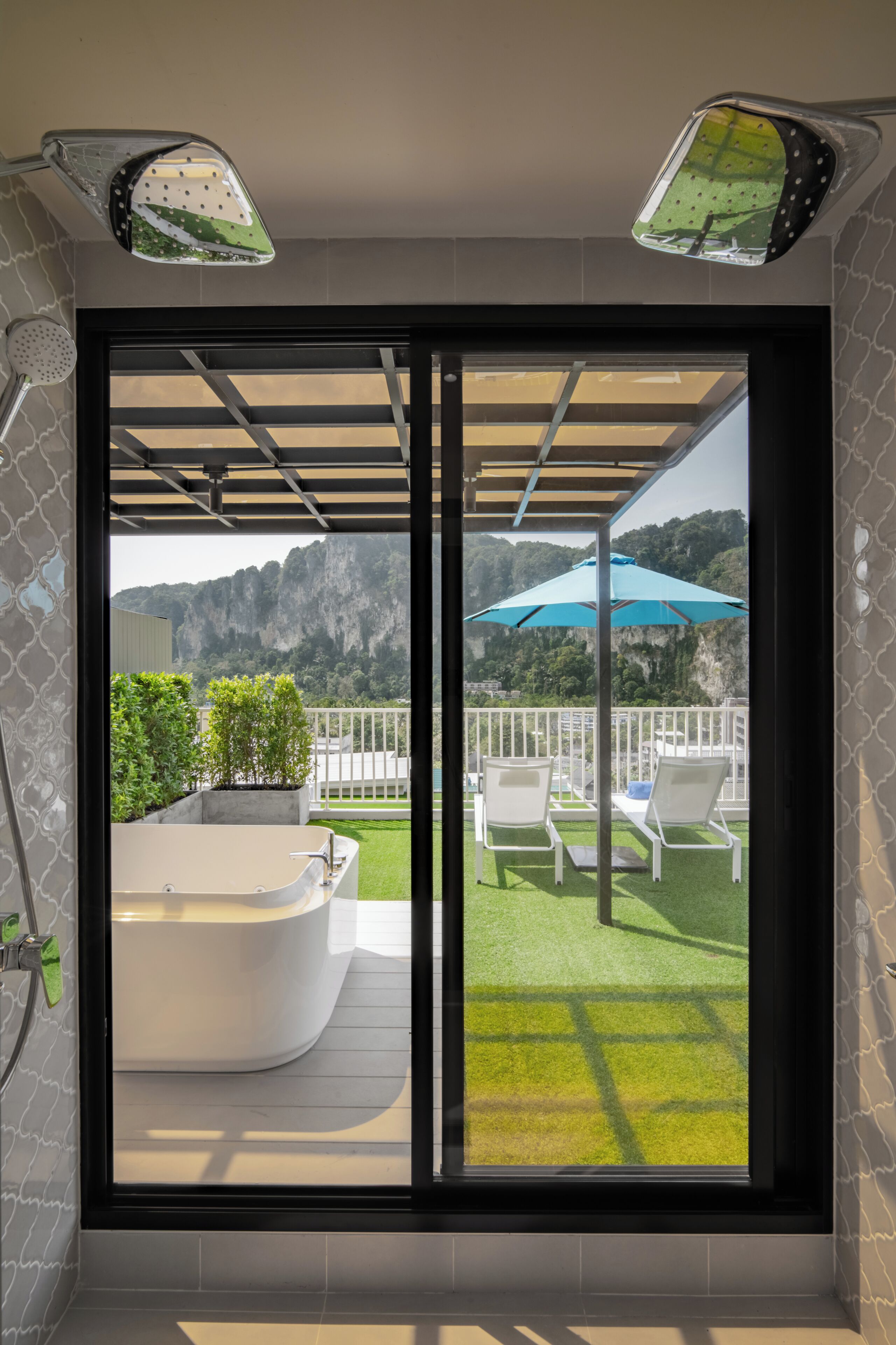 A bathroom with a bathtub, a shower, and a window with a view of a balcony with chairs and a blue umbrella.
