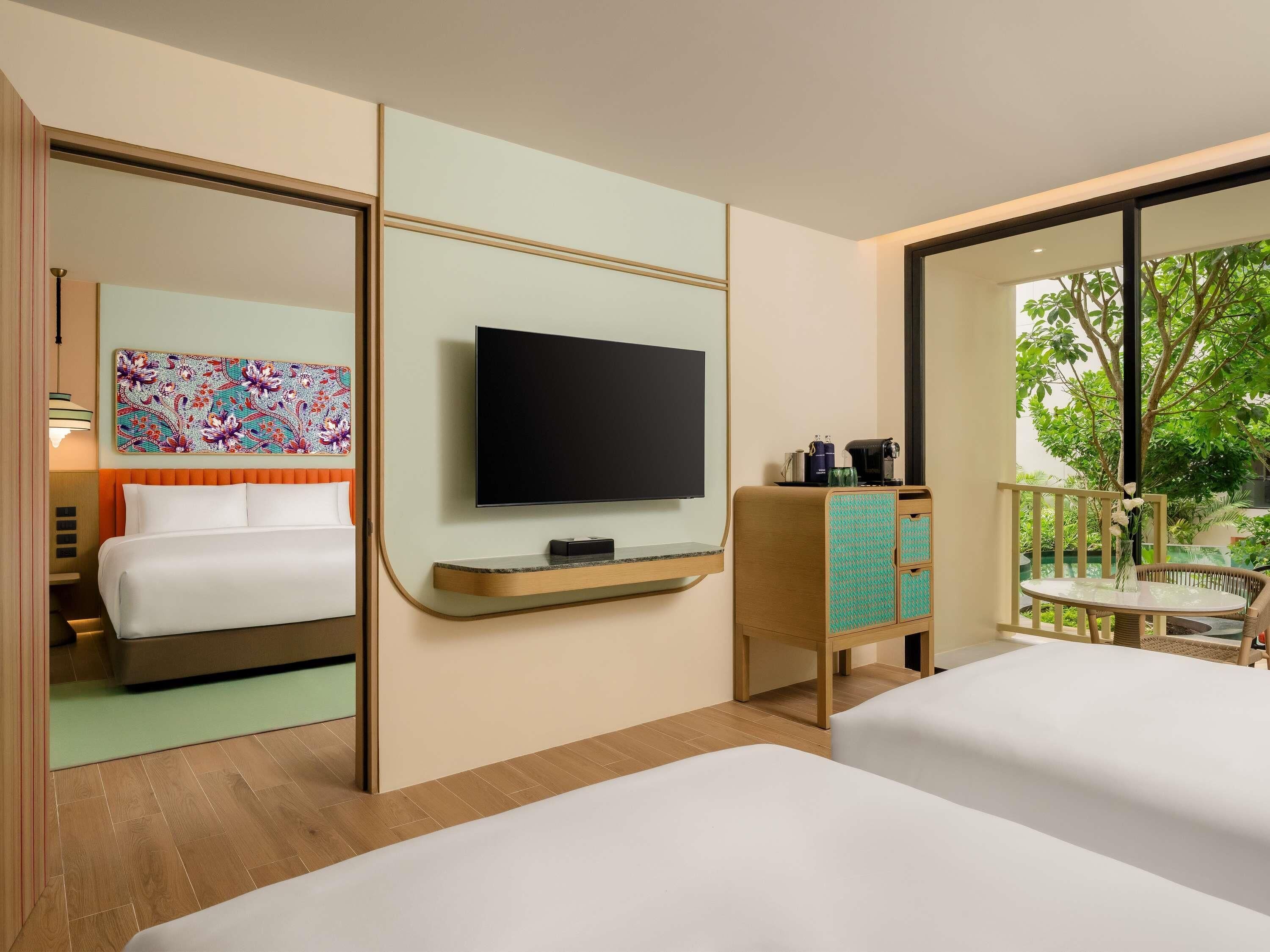 A hotel room with a bed, a TV, a small table, a cabinet, and a balcony with a view of trees.