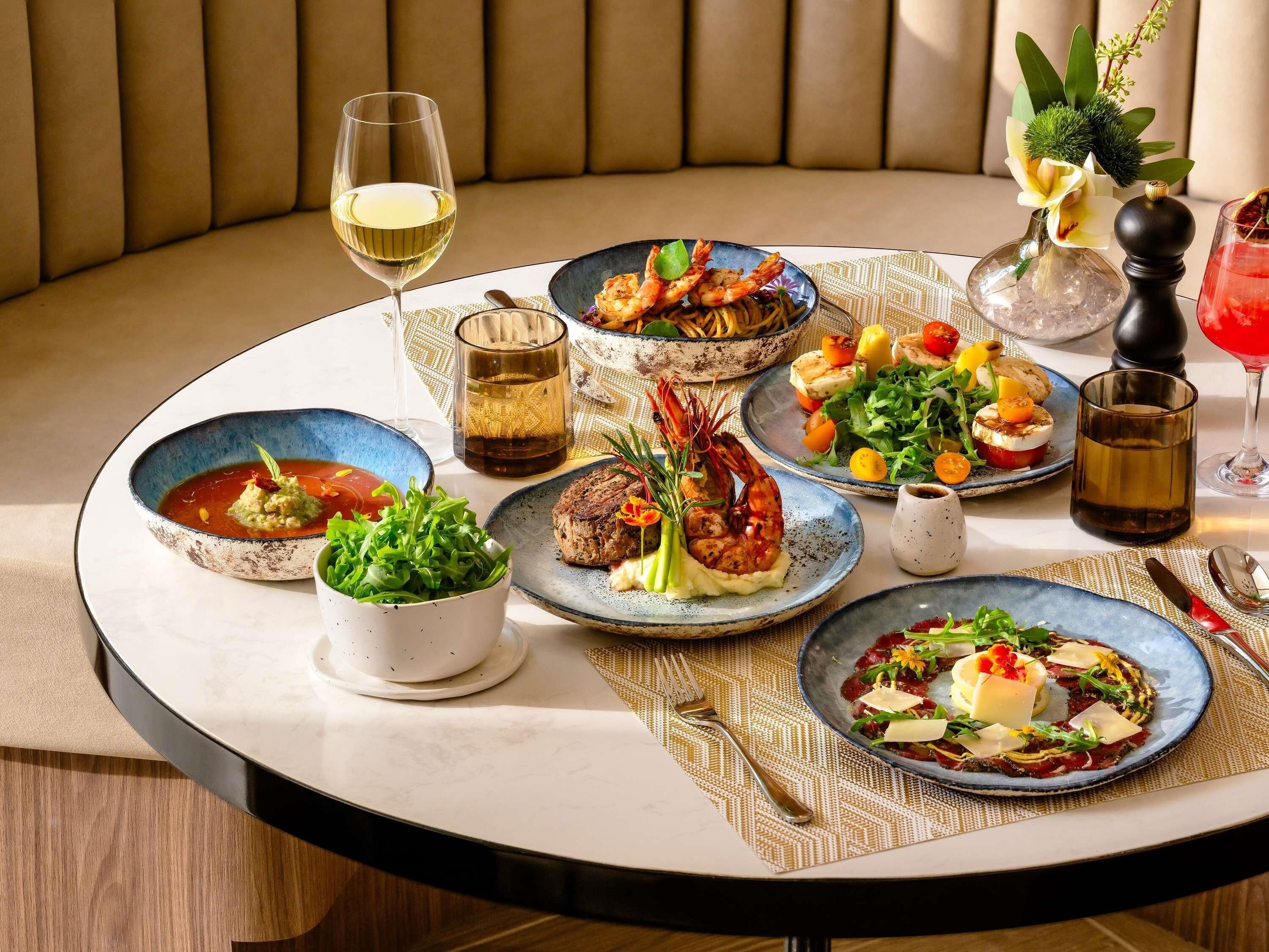 Una mesa redonda de comedor con diversos platos, una copa de vino blanco y un jarrón decorativo con flores.