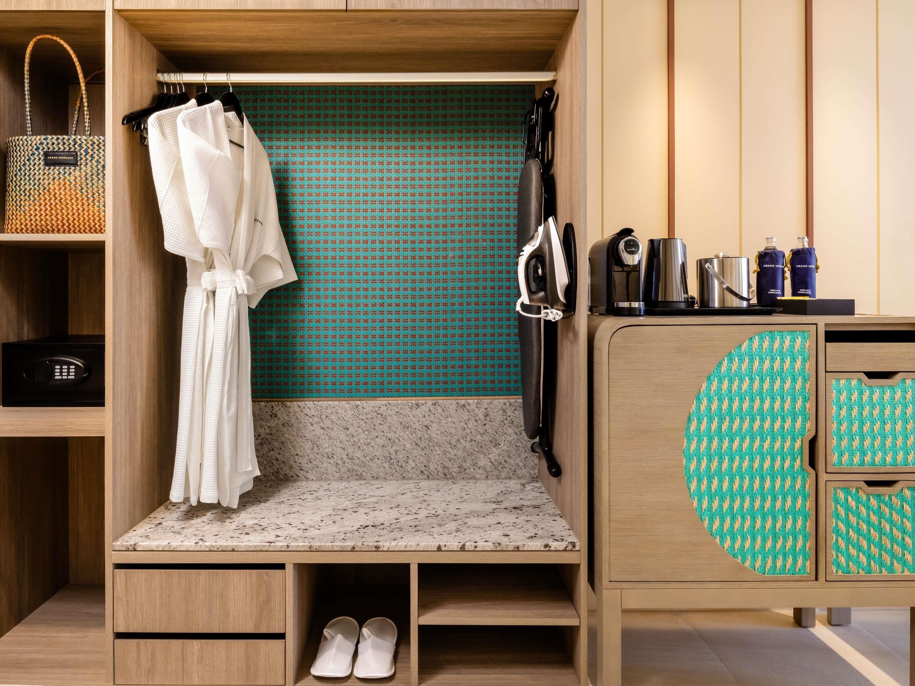 A compact hotel room with a kitchenette, a robe hanging, a pair of slippers, and a patterned cabinet.