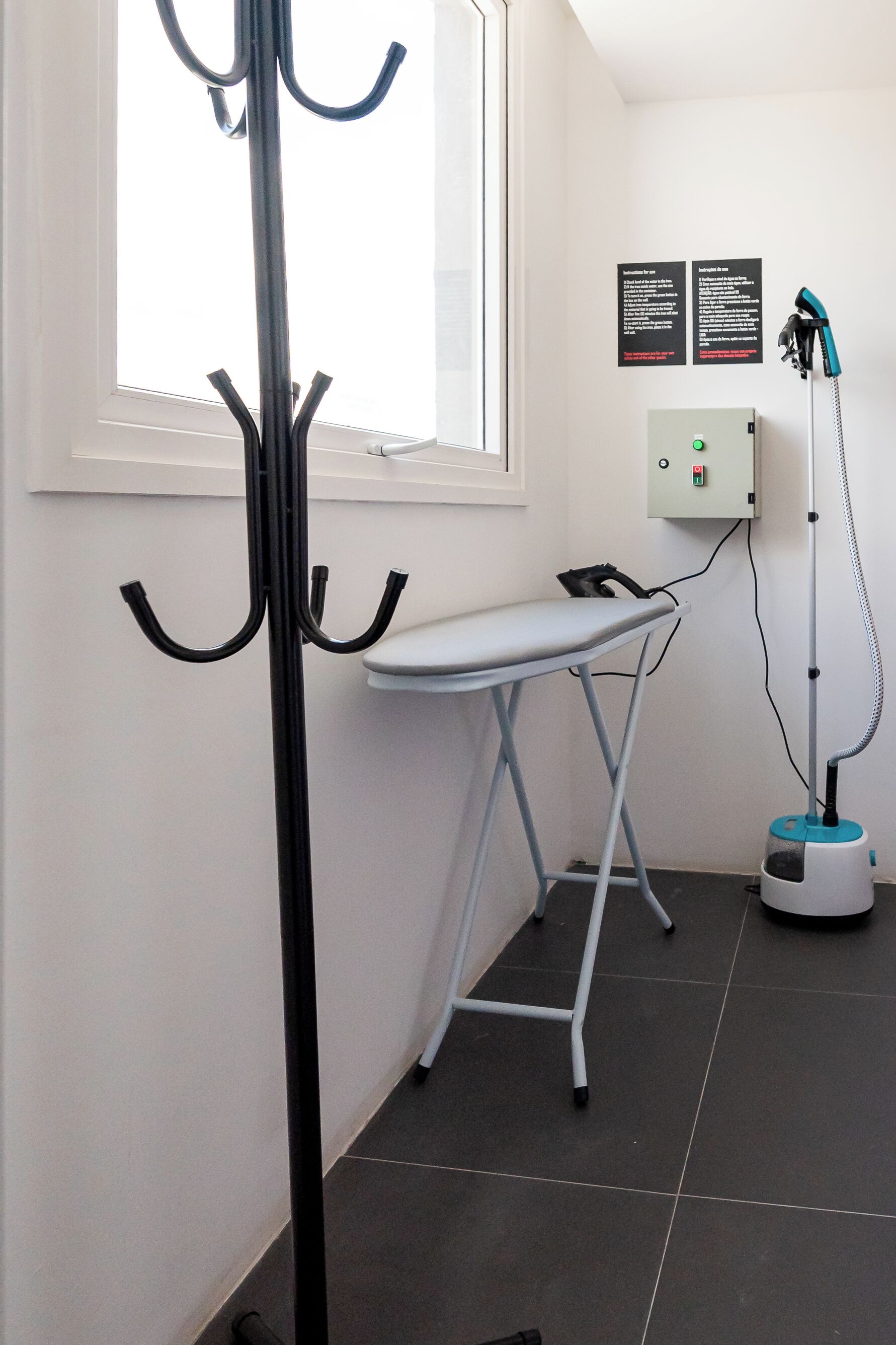 A room with a window, a coat rack, an ironing board, a vacuum cleaner, and two informational posters on the wall.