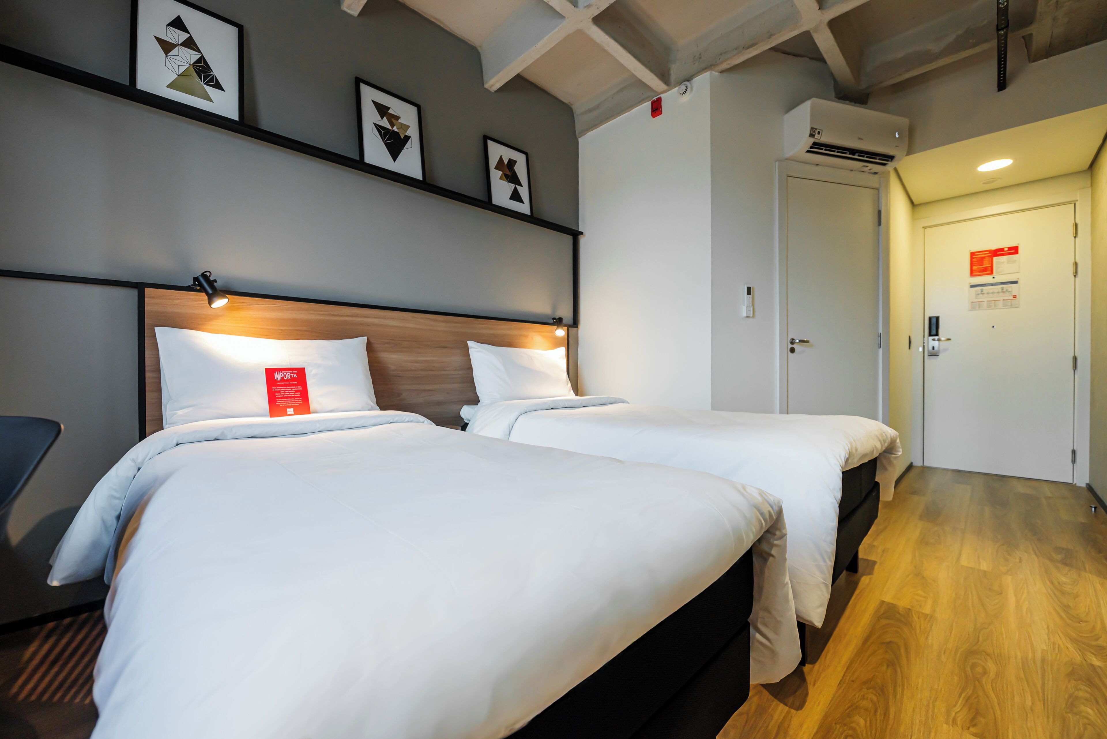 A hotel room with two beds, a wooden headboard, a wall-mounted air conditioner, and a door with a red sign.