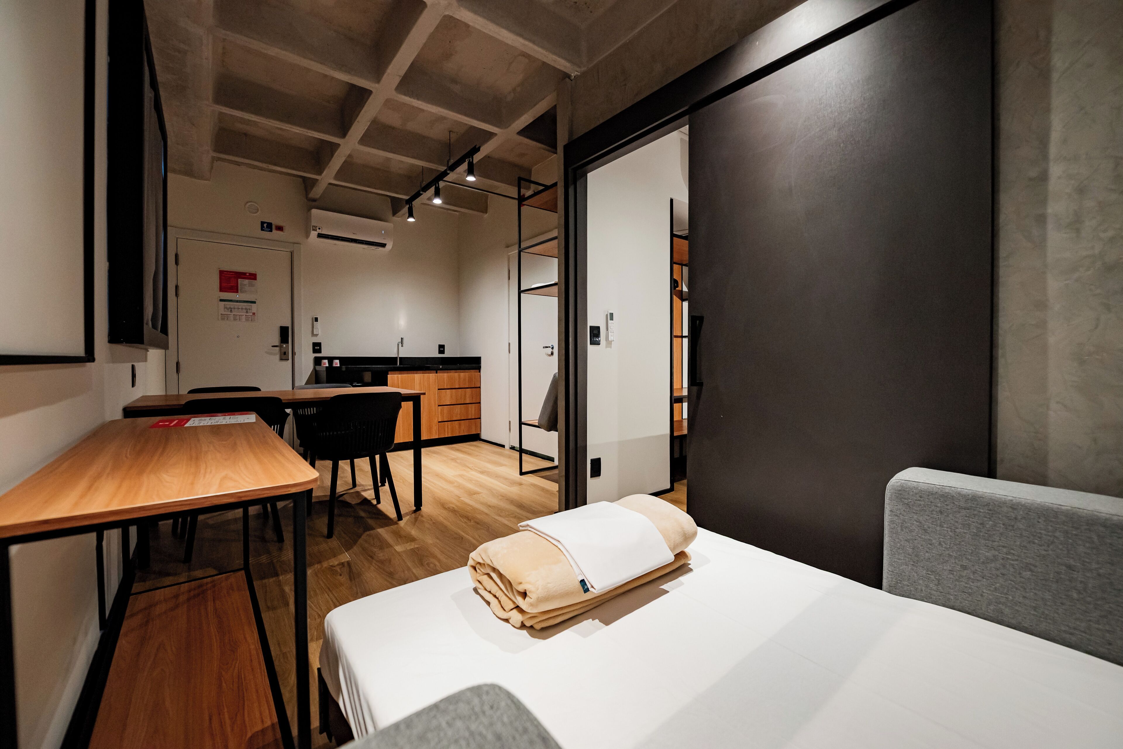 A modern hotel room with a wooden desk, a bed with a beige blanket, a grey couch, and a kitchen area in the background.