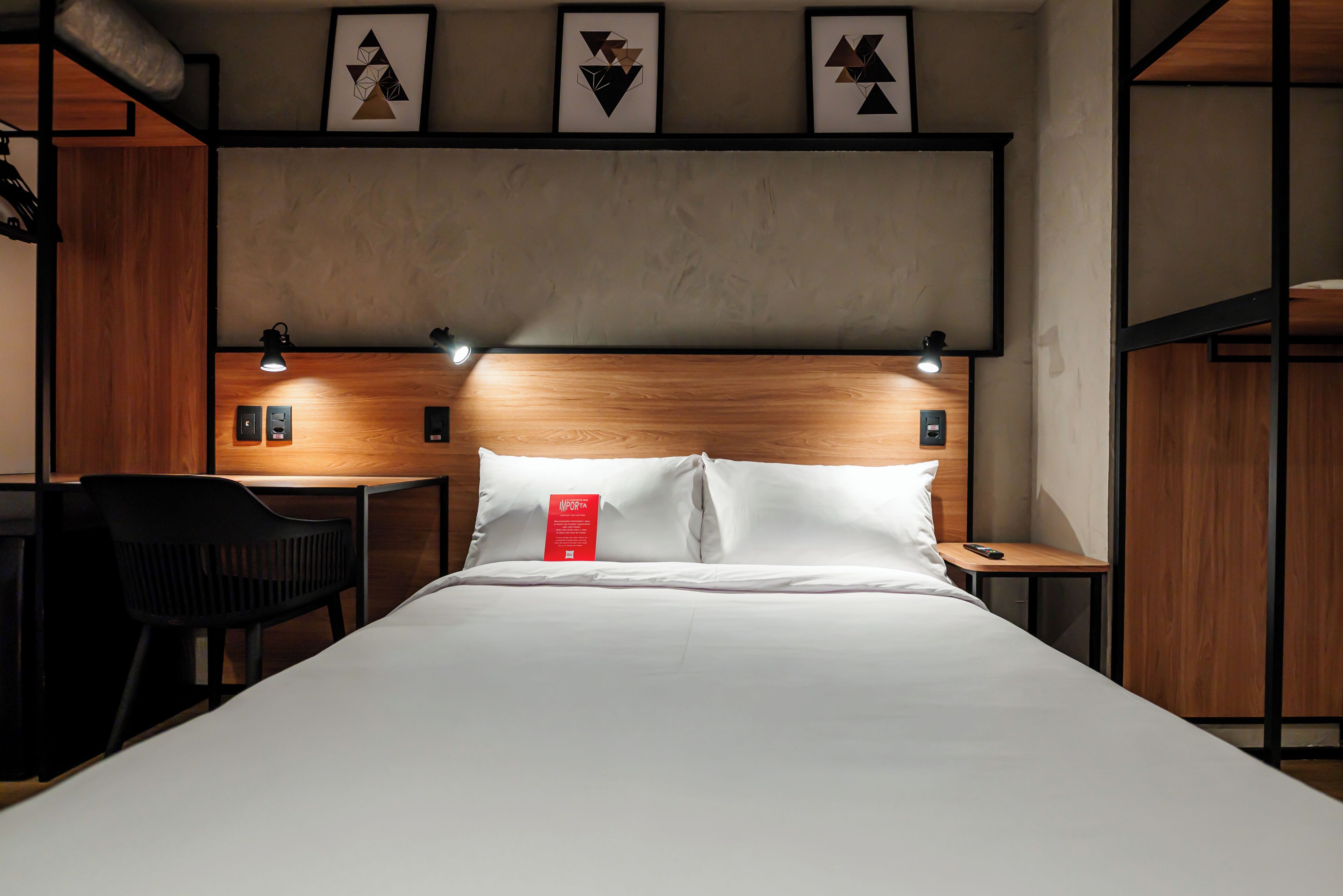 A hotel room with a wooden headboard, a bed with white linens, a desk with a chair, and framed artwork on the wall.