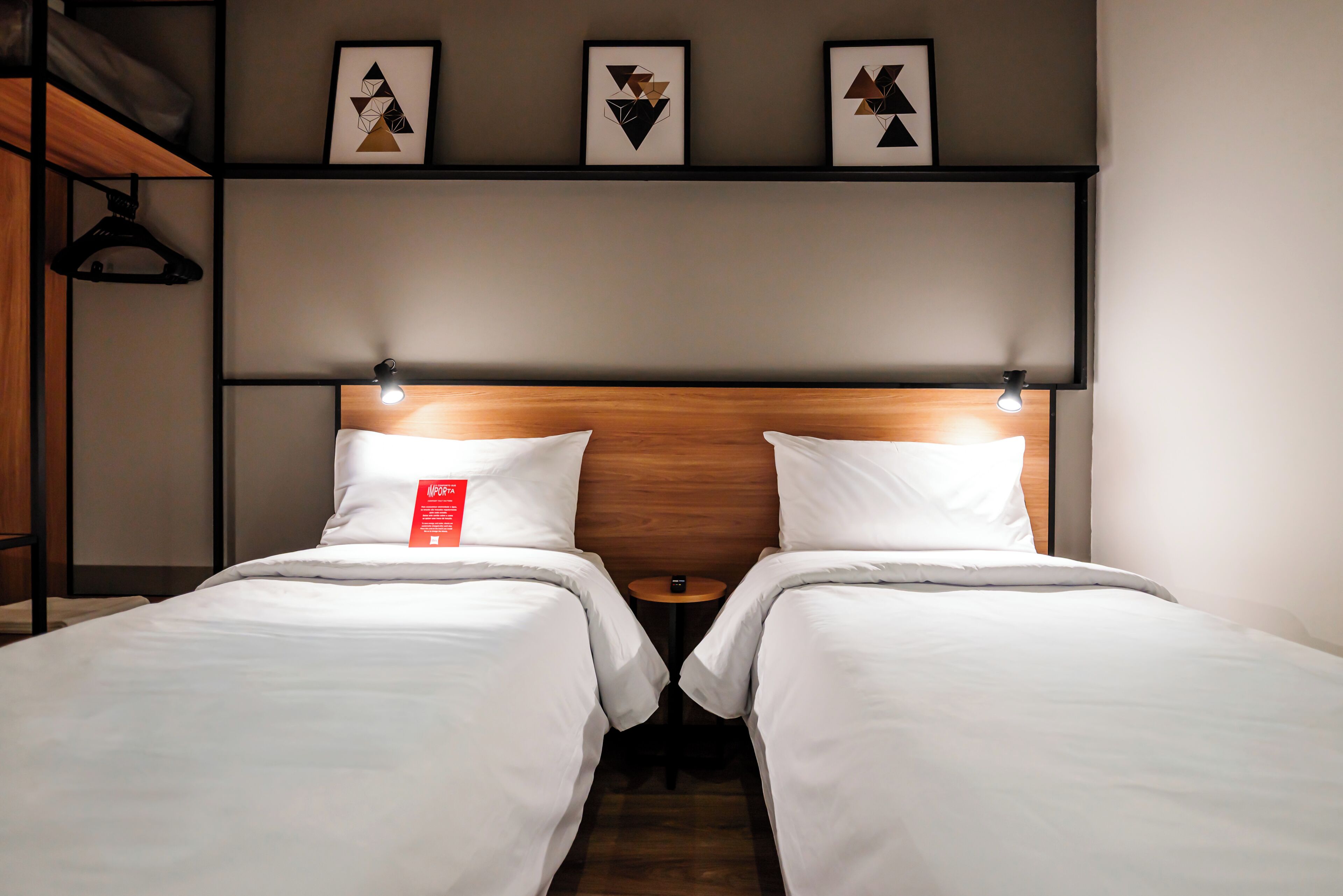 Habitación de hotel con dos camas, cabecera de madera y estante con obras de arte enmarcadas.