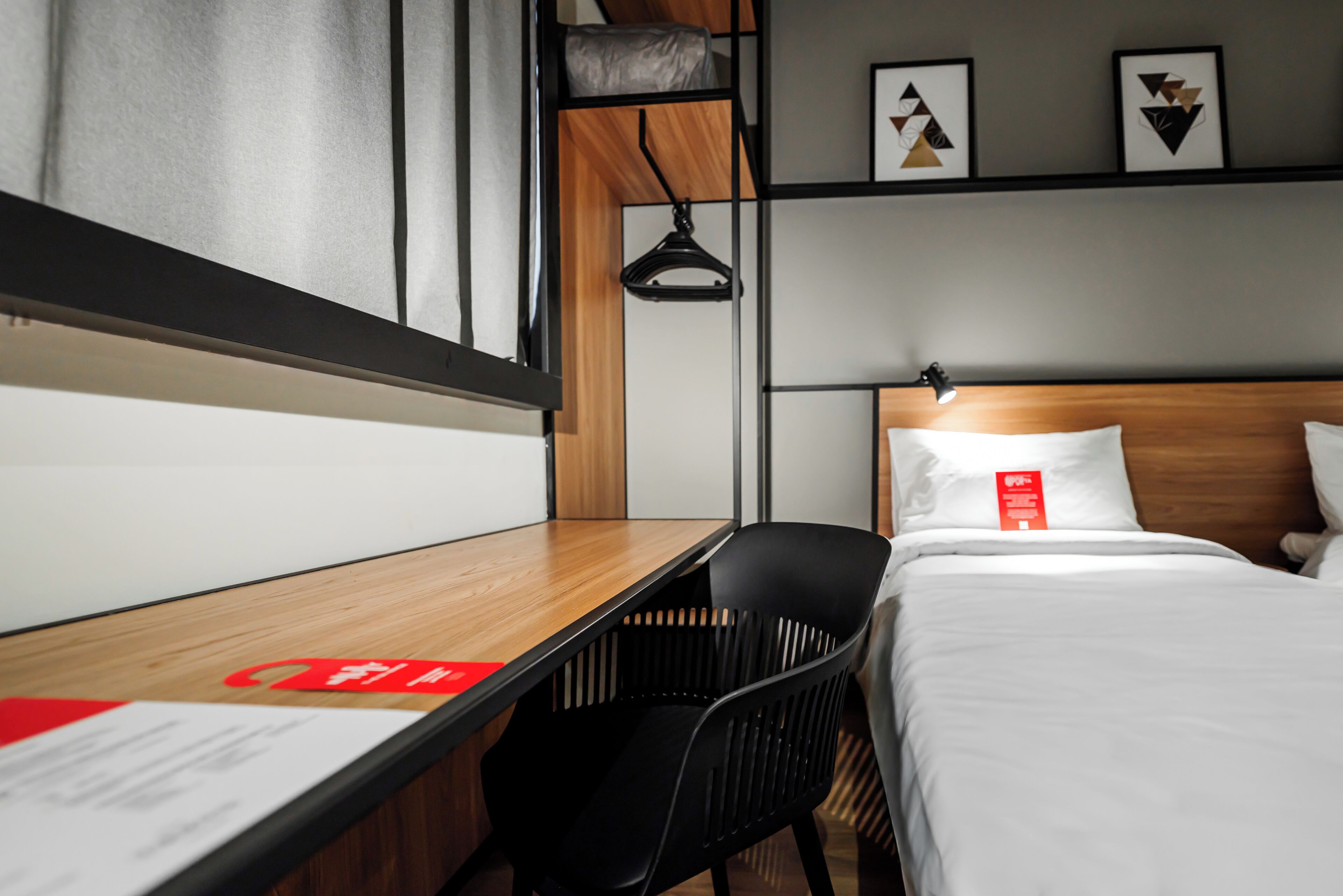 A hotel room with a wooden desk, a black chair, a bed with white bedding, a hanging light, and abstract wall art.