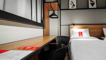 A hotel room with a wooden desk, a black chair, a bed with white bedding, a hanging light, and abstract wall art.