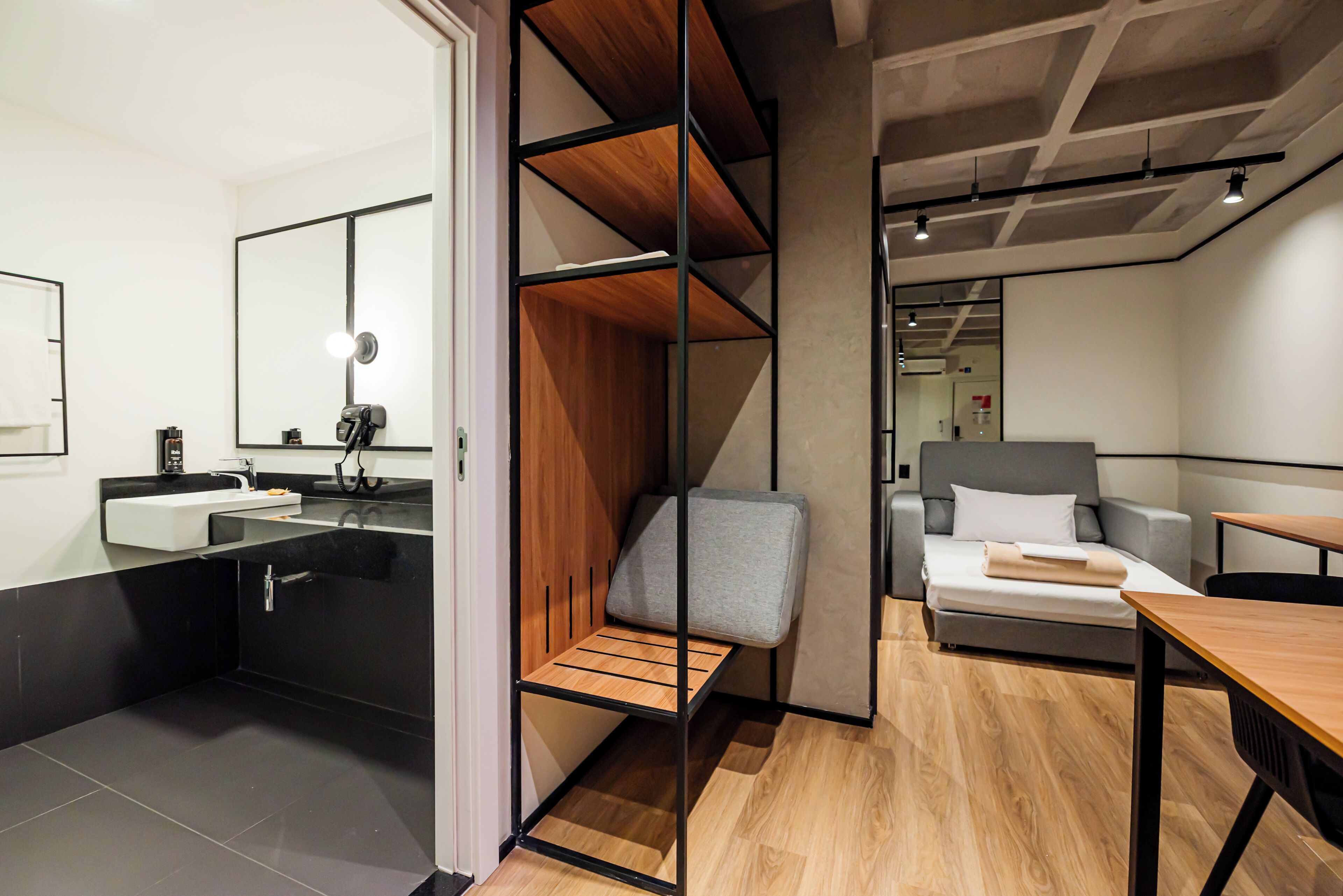 A modern hotel room with a wooden bench, a grey sofa, a bed, and a desk.