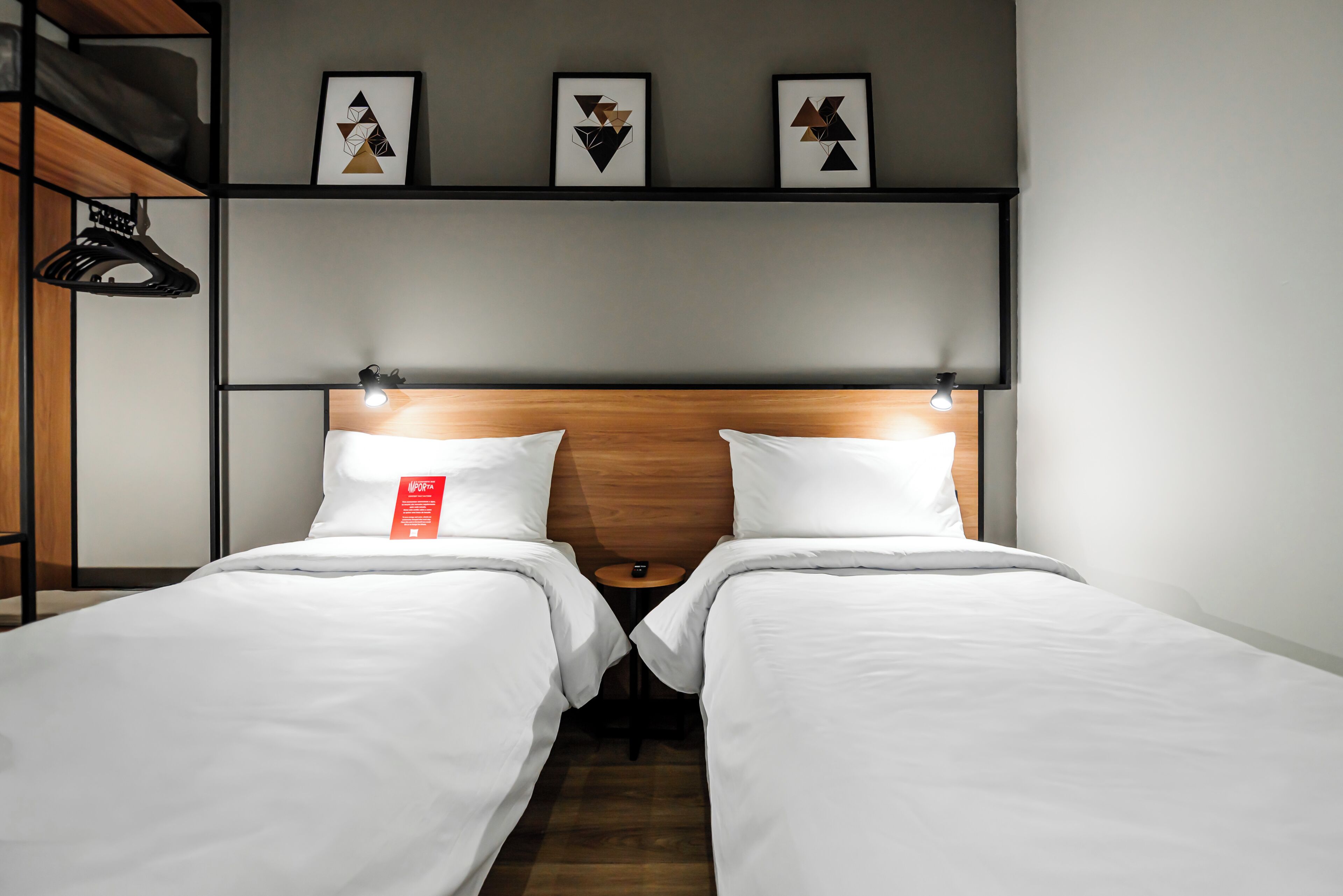 Quarto de hotel com duas camas, cabeceira de madeira, luminária na parede e três quadros pendurados acima das camas.
