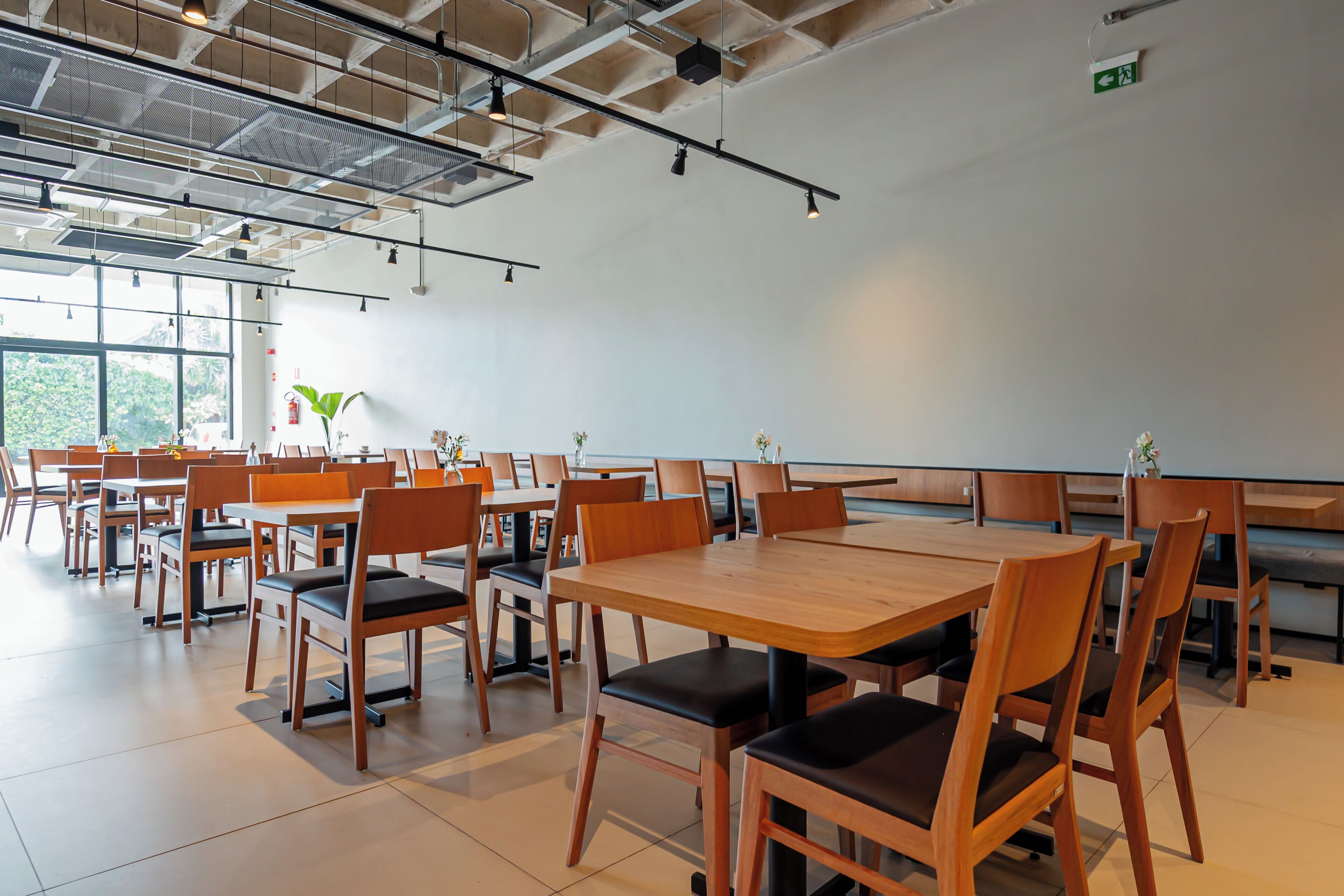 A spacious dining area with multiple wooden tables and chairs, large windows, and a high ceiling with exposed beams.