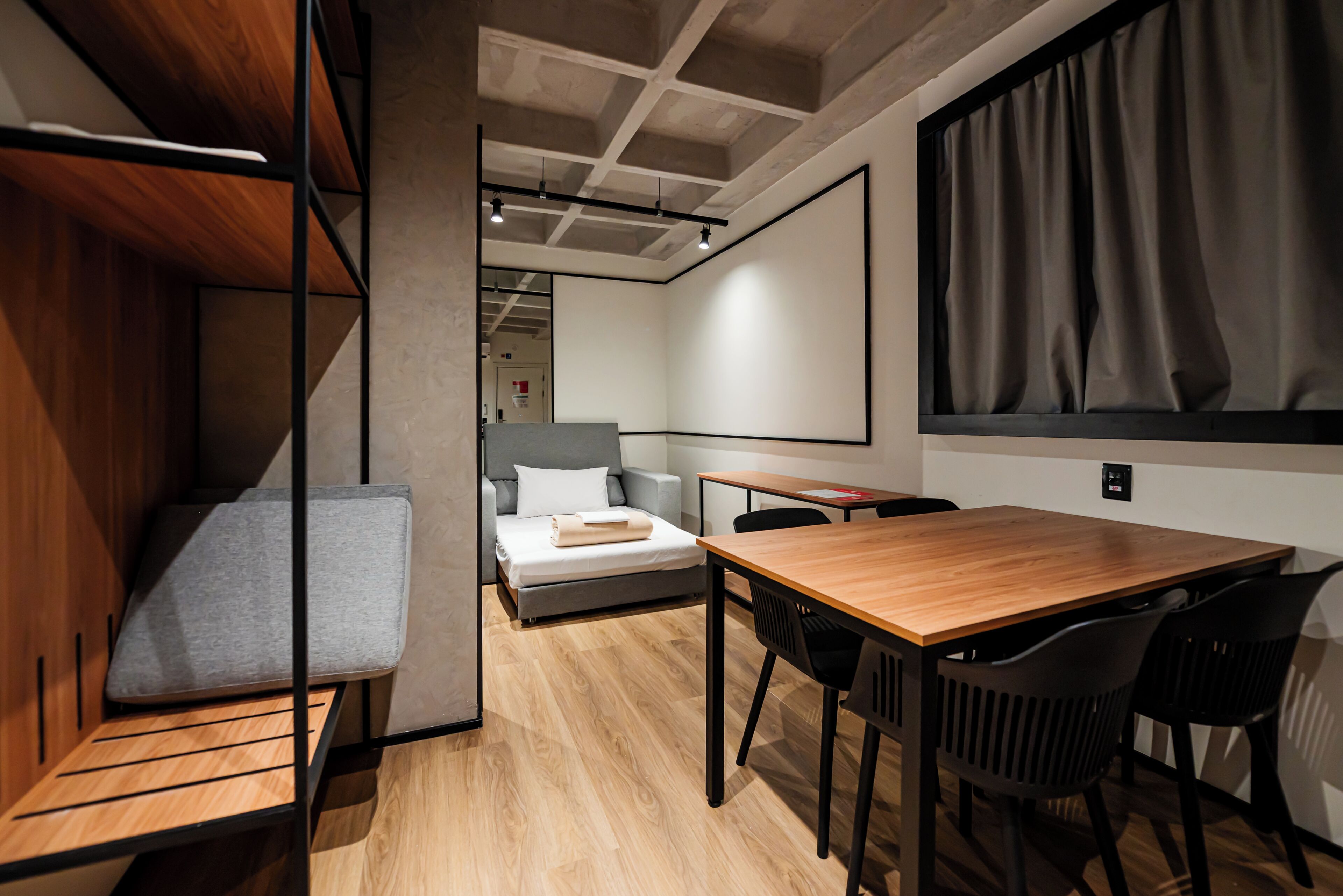 Una habitación de hotel moderna con un escritorio de madera, un sofá gris, una cama y un amplio ventanal con cortinas.